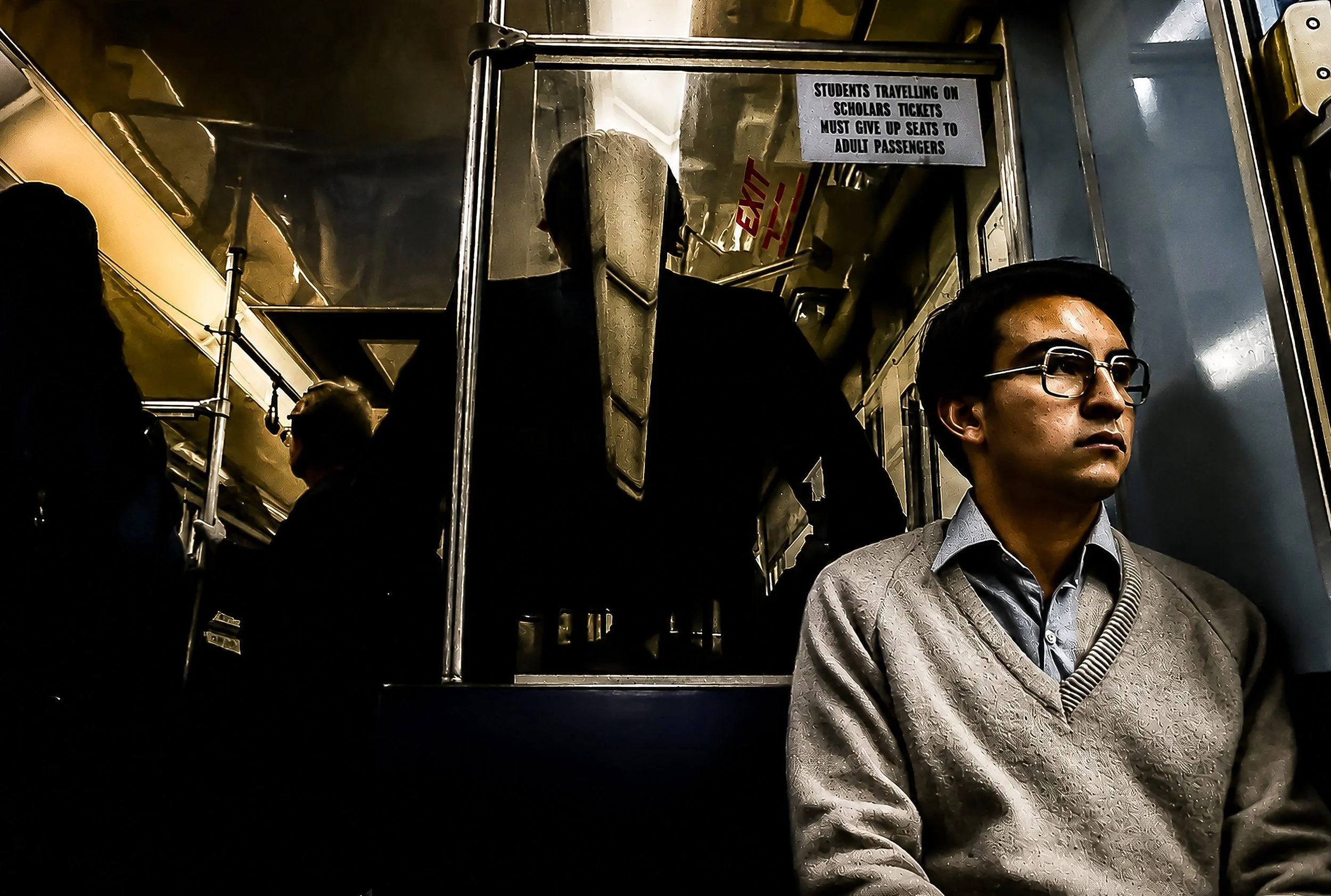 A man with glasses and a gray sweater standing inside a bus, with other passengers and metal handrails visible around him. There is a sign that reads 'Students traveling on scholars tickets must give up seats to adult passengers.'
