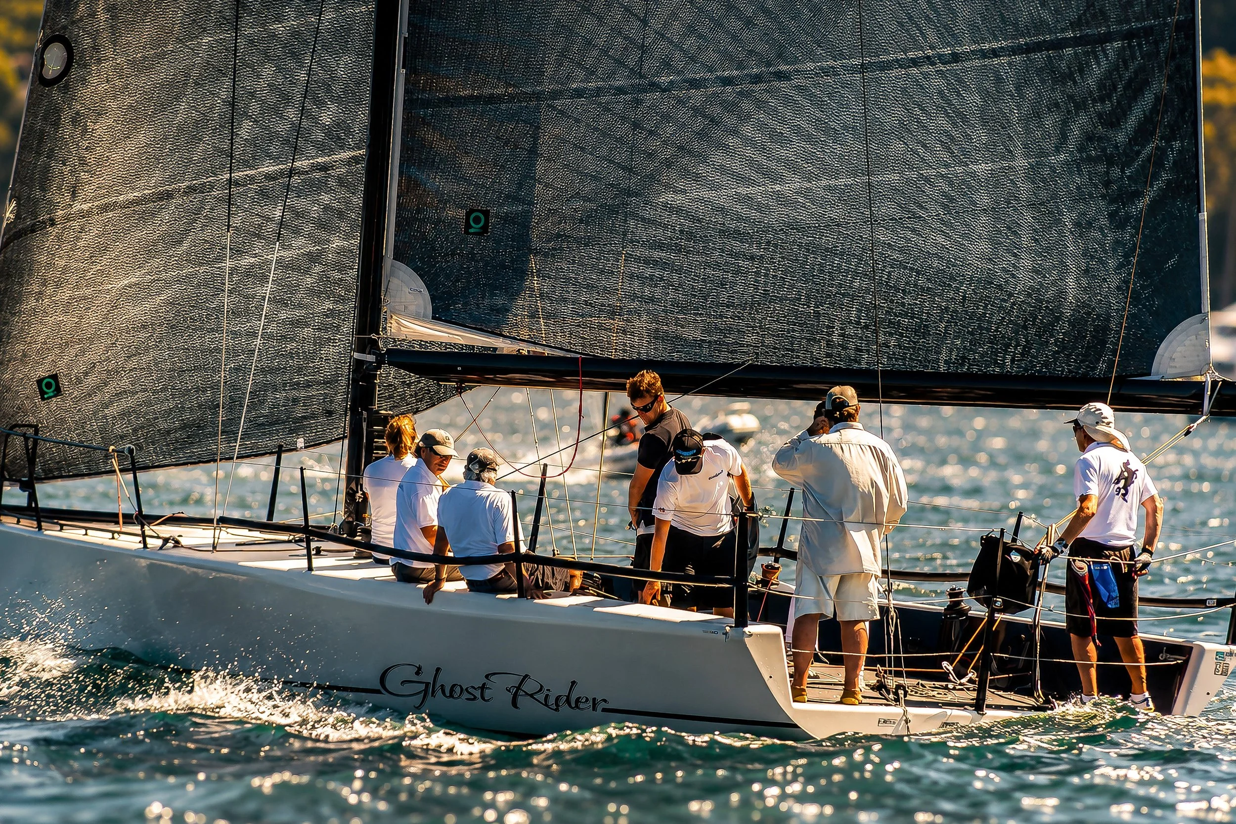 A sailboat on the water with a crew of people dressed in white shirts and hats, the boat named Ghost Rider, with large black sails, during a sunny day.