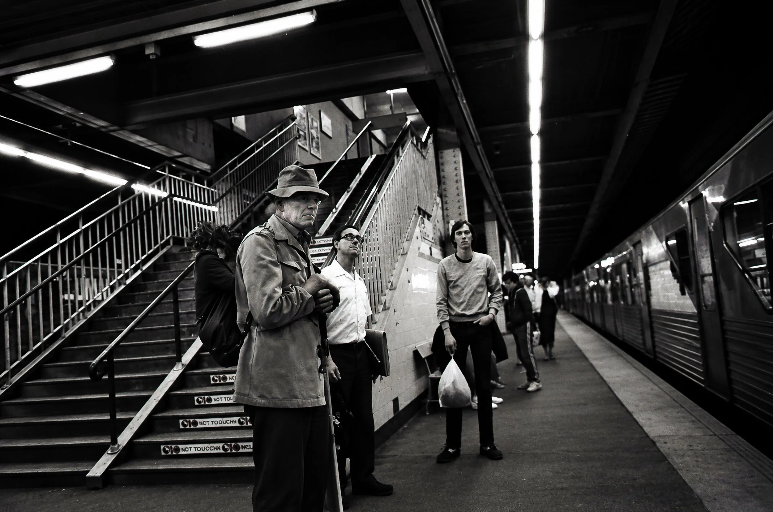 Down the Platform - Passengers move through a darker section of the station beside the railway tracks. The stairs, ironwork, fluorescent lighting and train create a strong sense of depth and movement beneath the city.