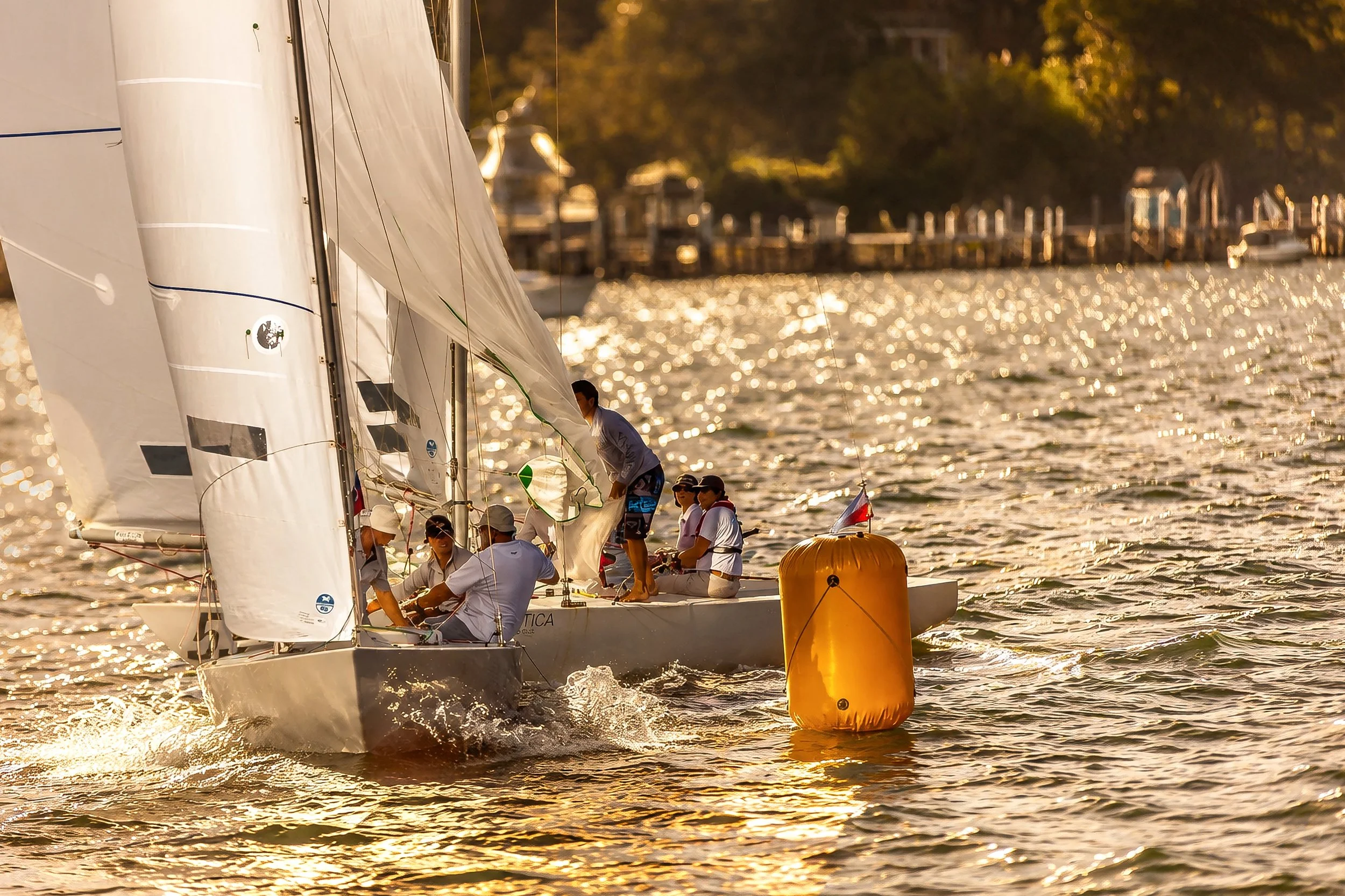 Sailboats racing around a buoy on a sunny waterway, with multiple team members on each boat working together, and a background of trees and docks.