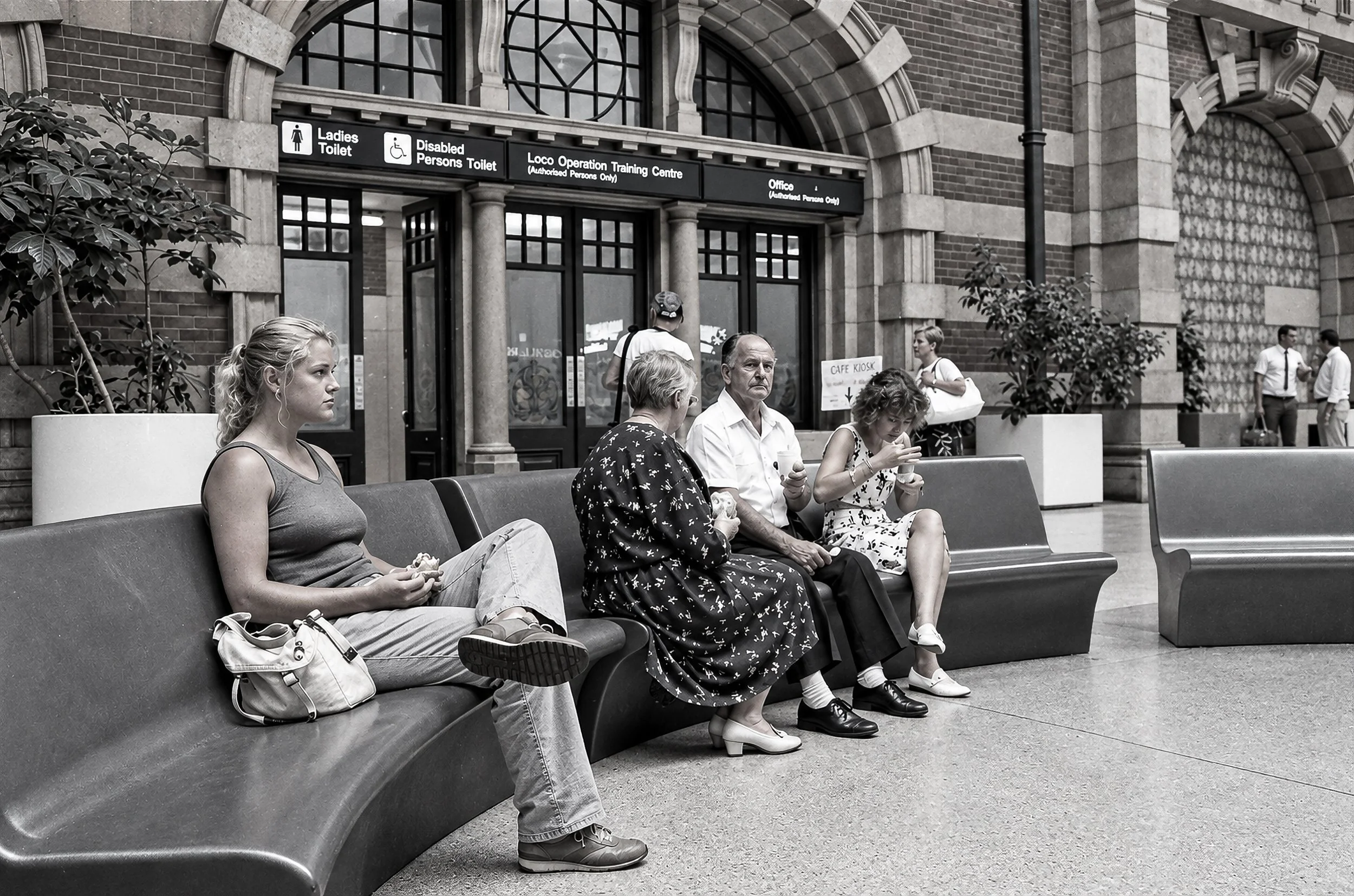 Waiting at the Station - Passengers sit beneath the grand architectural details of the station concourse. The high arches, brickwork, signage, curved seating and everyday activity combine to create a broader portrait of Sydney's railway world in the 