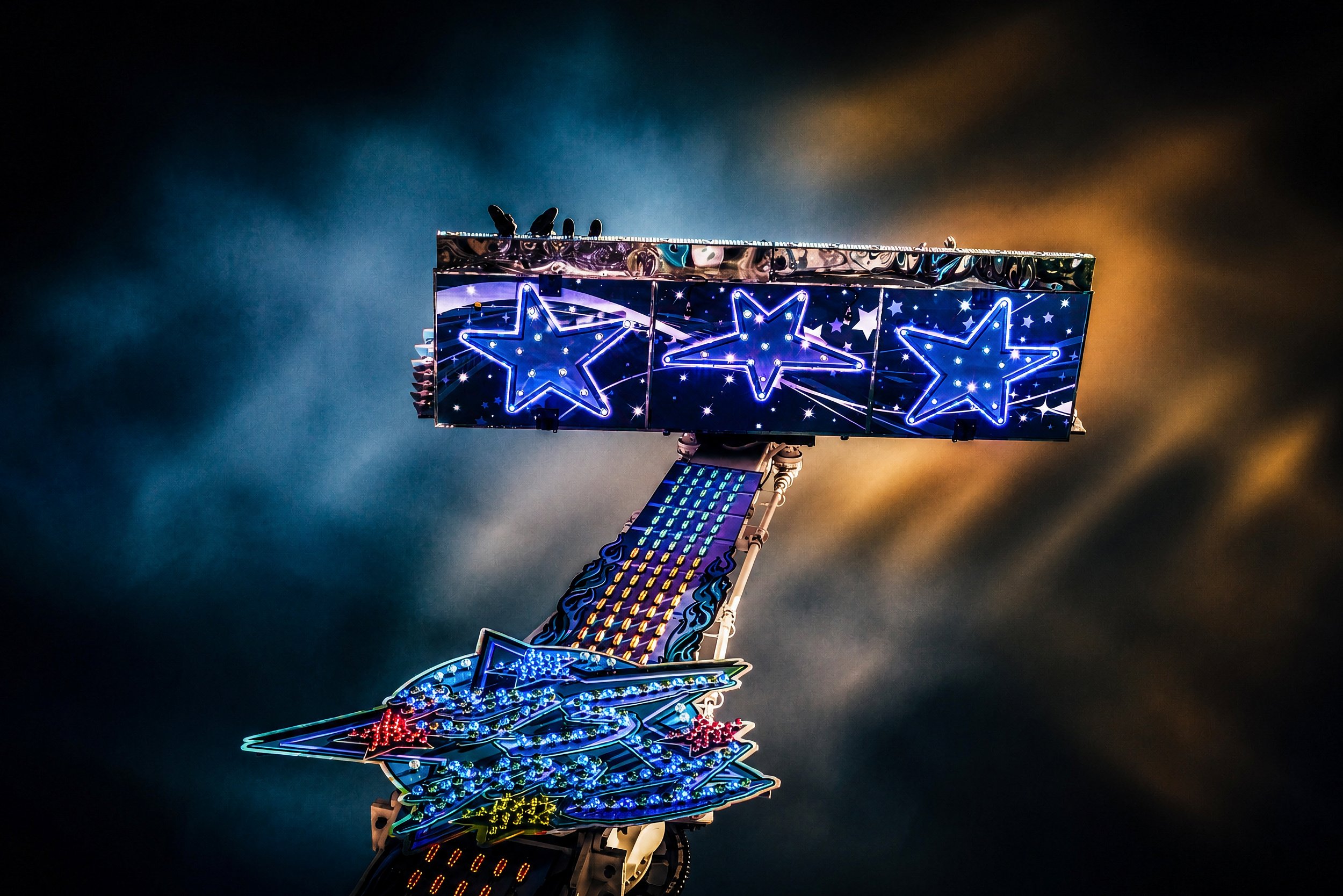An illuminated carnival ride decorated with glowing blue stars photographed against a dramatic night sky.
