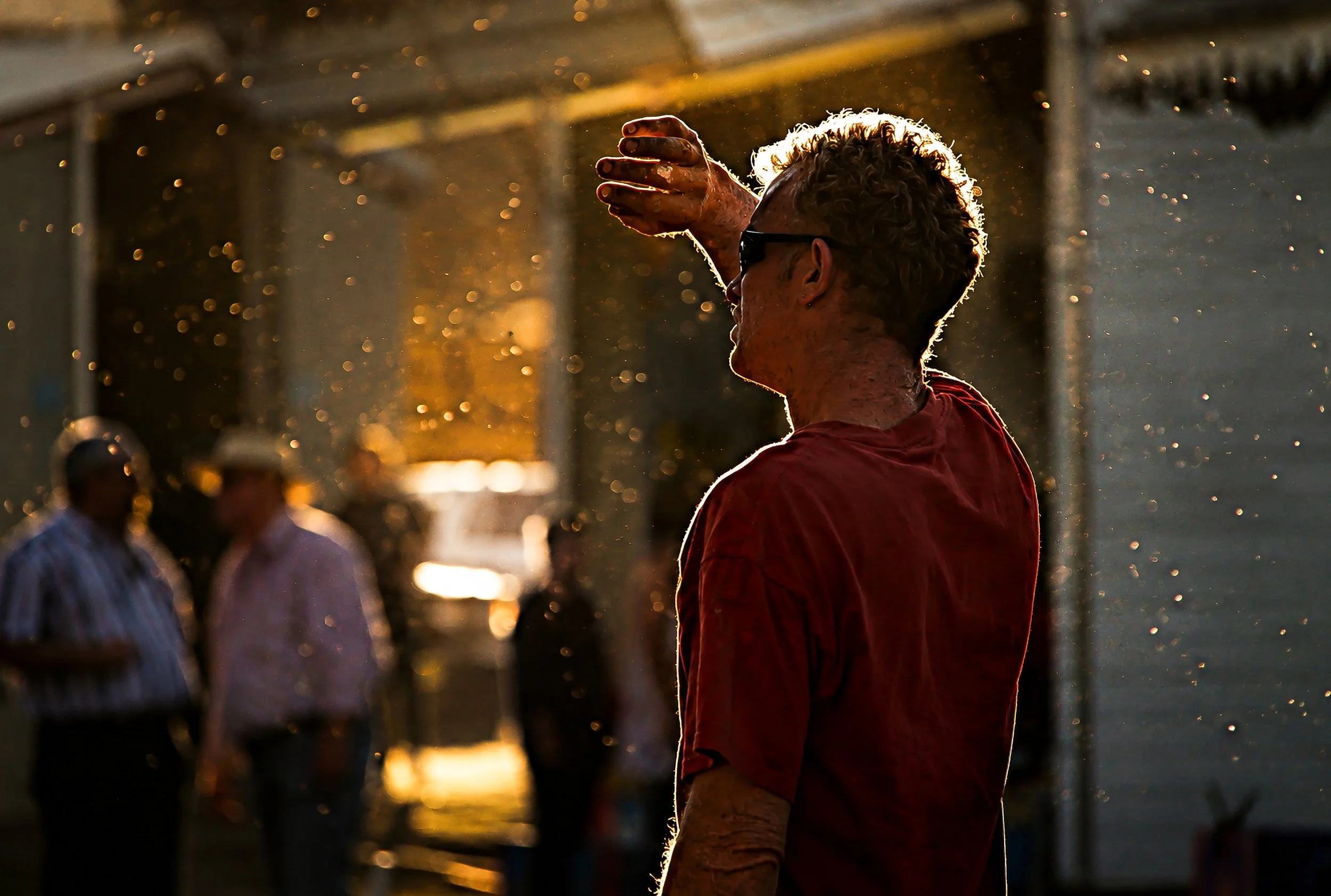 A person with curly hair and sunglasses, wearing a red shirt, holding their hand up with fingers extended, with a city street scene and sunlight in the background, creating a silhouette effect.