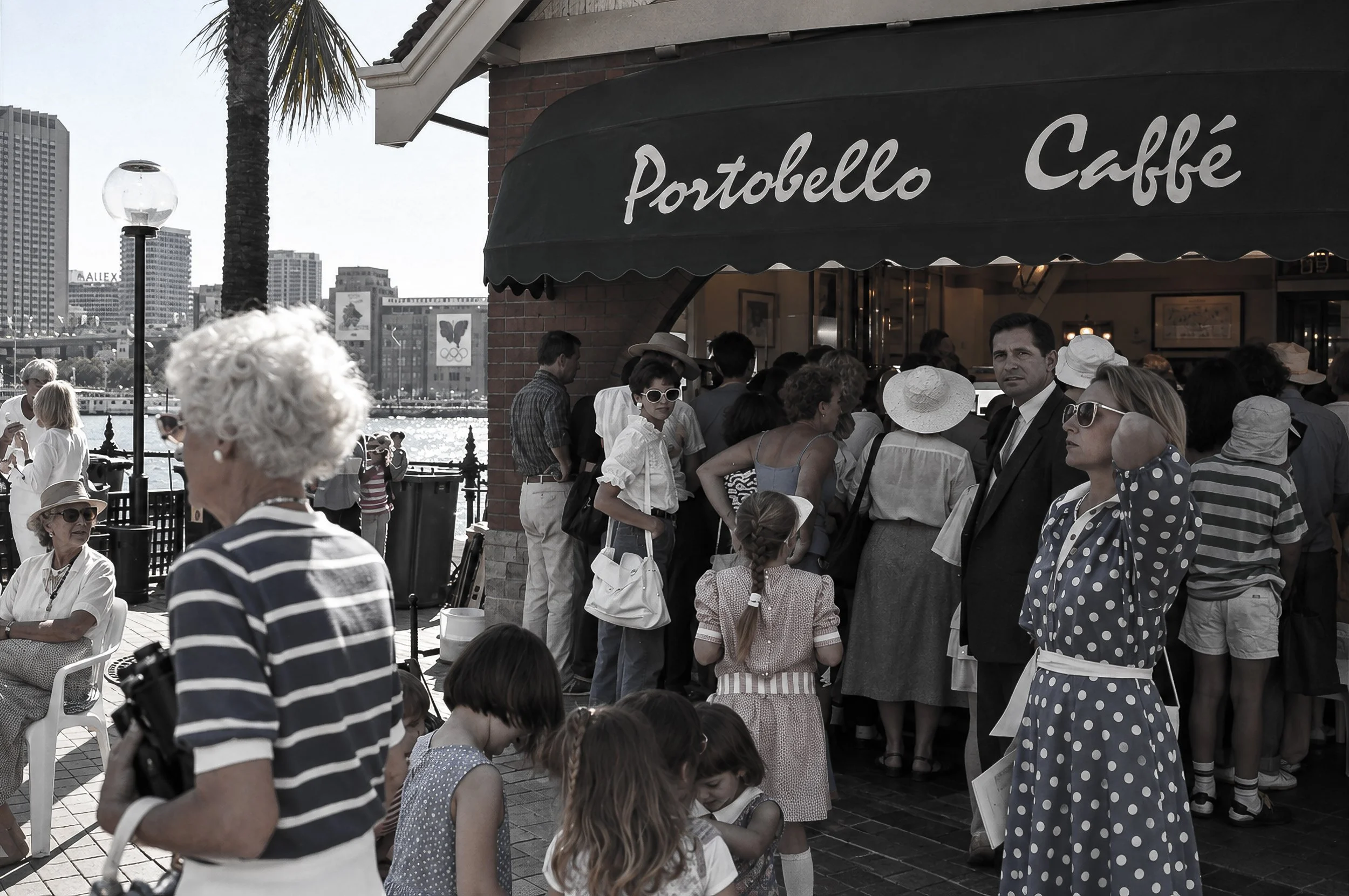 Portobello Caffé - Sydney Waterfront -People gather outside Portobello Caffé beside Sydney Harbour, creating a lively scene of conversation, movement and waterfront activity. The café signage and surrounding architecture anchor the photograph firmly 