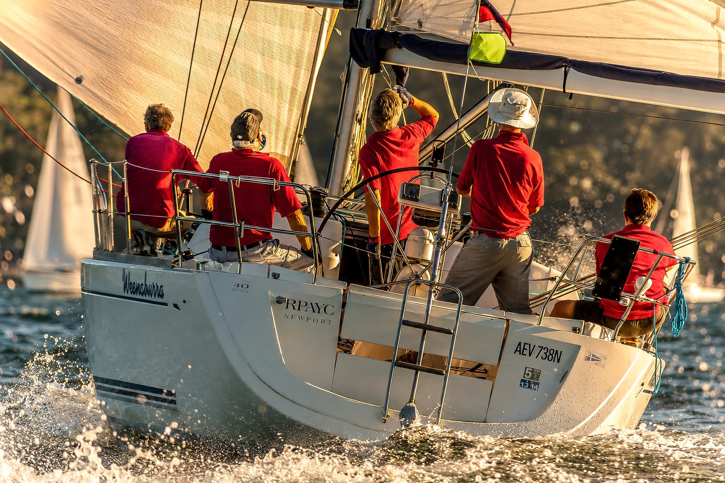 A group of five people wearing red shirts sailing on a white sailboat during sunset, with a large sail catching the wind and water splashing at the boat's bow.
