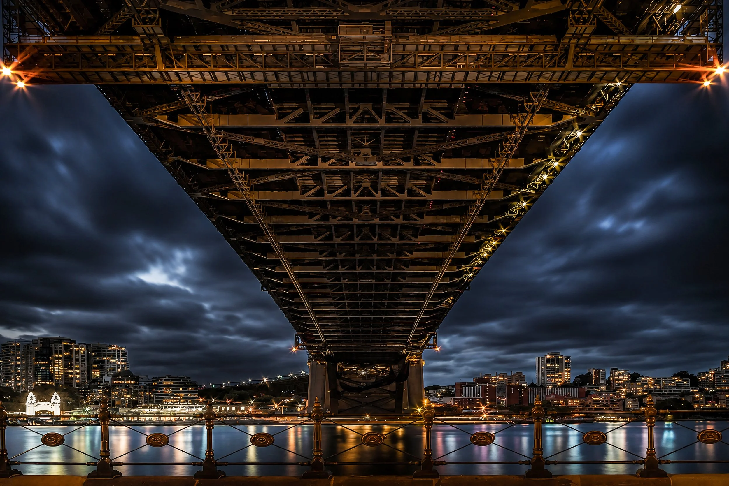 A dramatic symmetrical perspective beneath a major Sydney bridge at blue hour with city lights reflected on the water.
