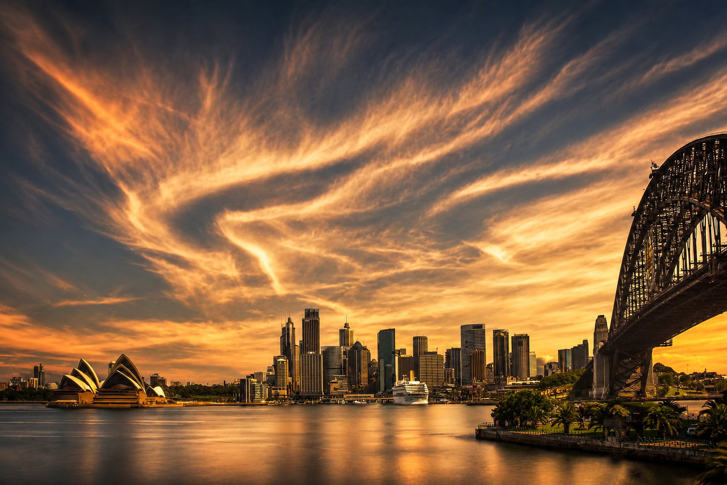 Sweeping sunset clouds arc above Sydney Harbour as the Harbour Bridge frames the city skyline in warm evening light.
