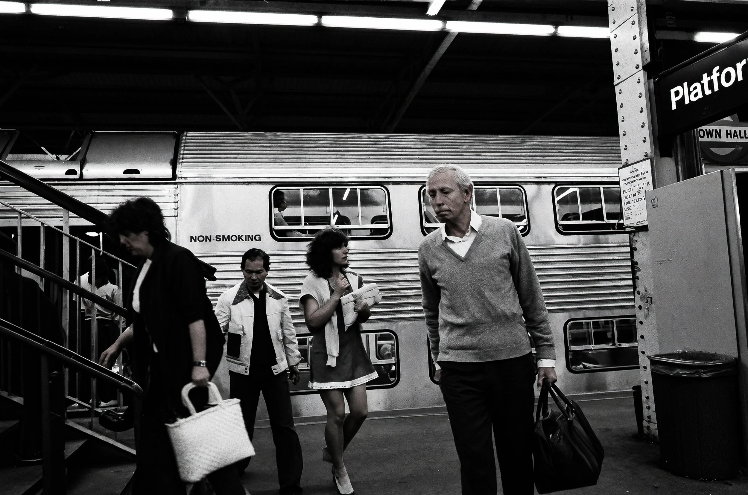Between Trains - Passengers move along the platform beside a waiting train. The contrasting clothing, expressions and directions of movement give the photograph the spontaneous quality of an unposed encounter.
