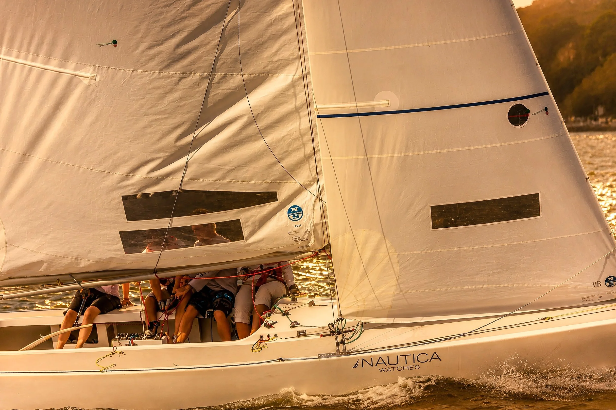 A sailboat with a white sail on a body of water during sunset, with people onboard, and trees visible in the background.