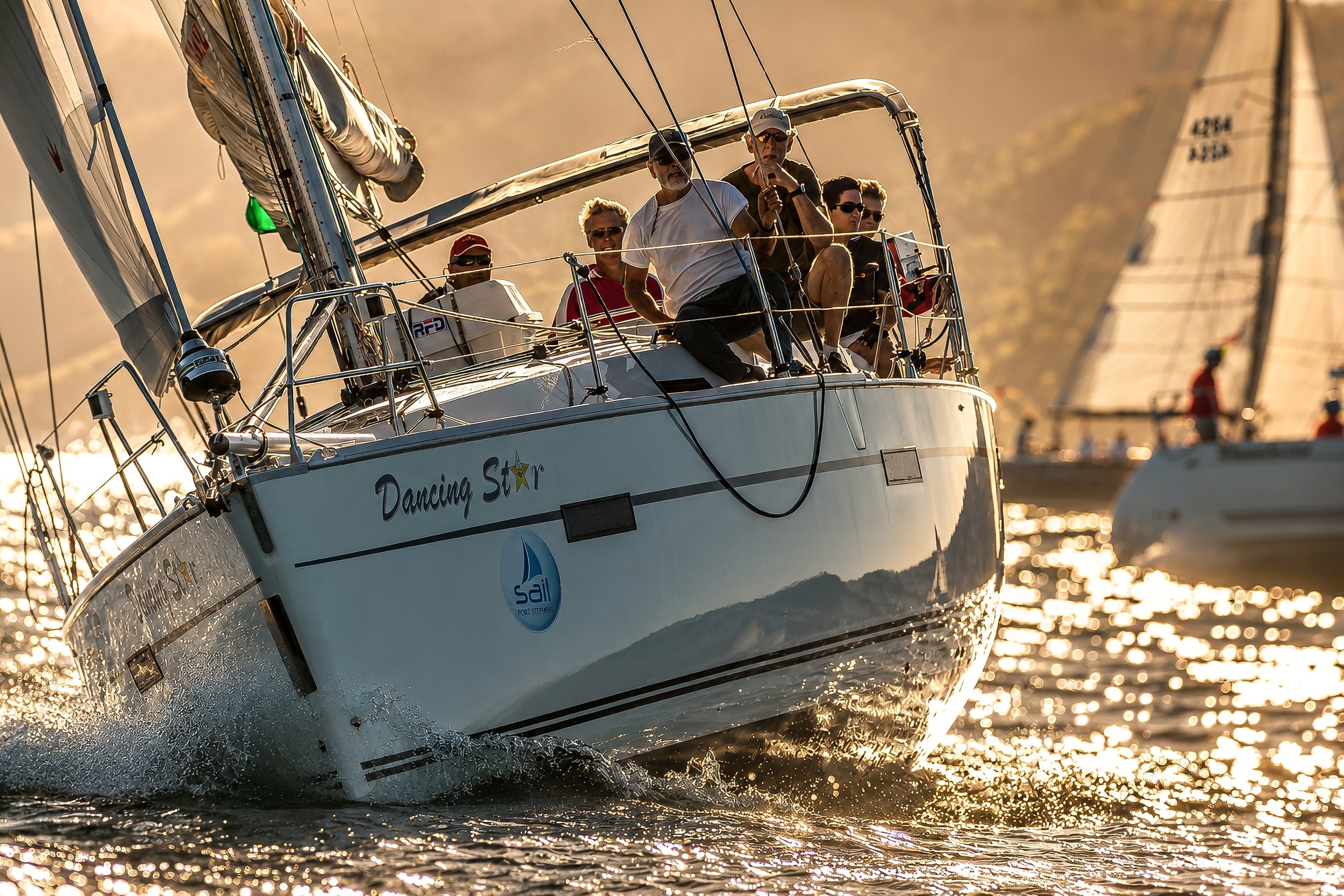 A group of people sailing on a boat named 'Dancing Star' during sunset, with several other sailboats visible in the background.