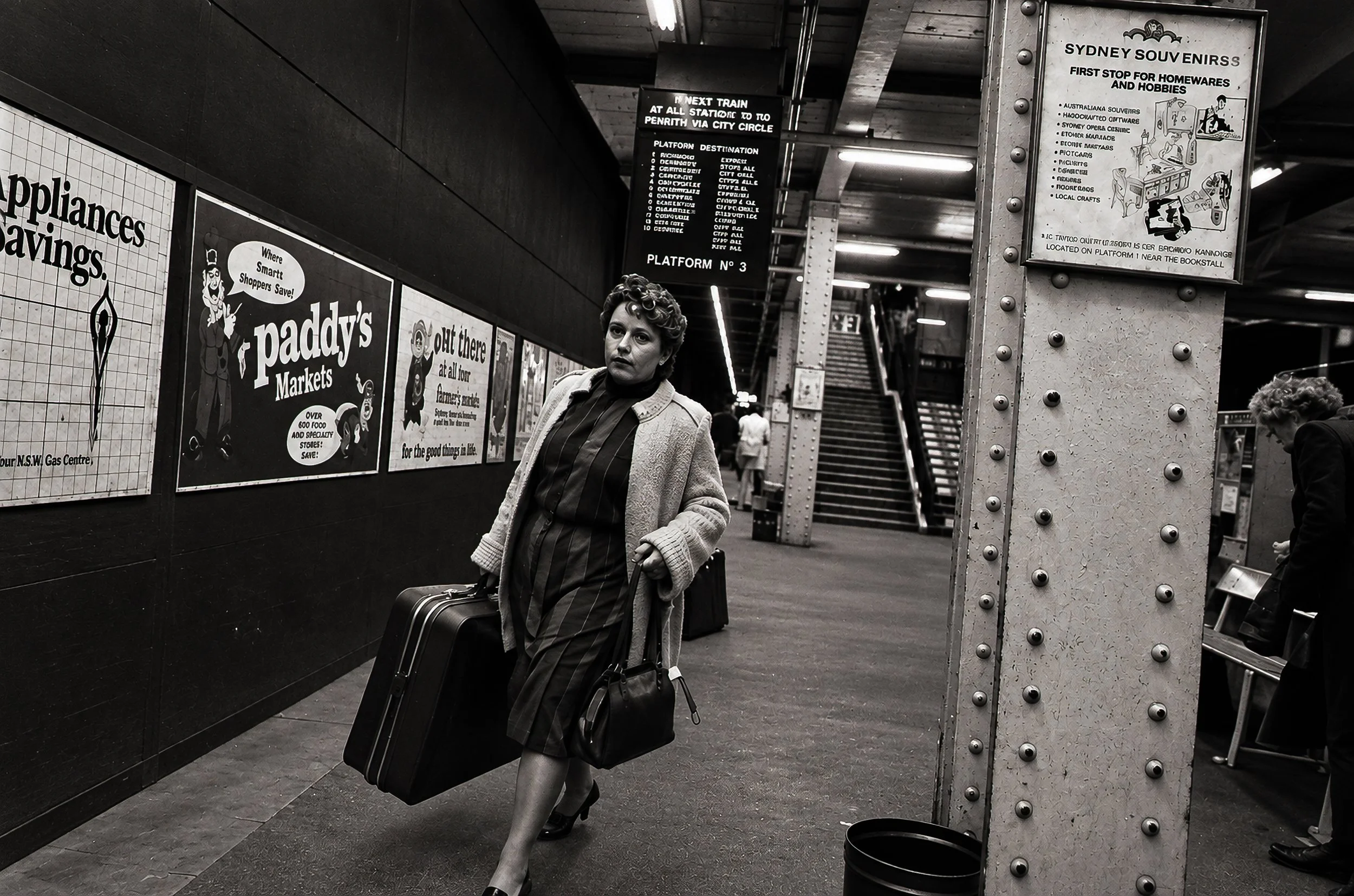 The Traveller - A woman carrying luggage walks along the platform beneath rows of period advertisements. The railway architecture and advertising provide a strong sense of place while her movement gives the photograph its narrative.