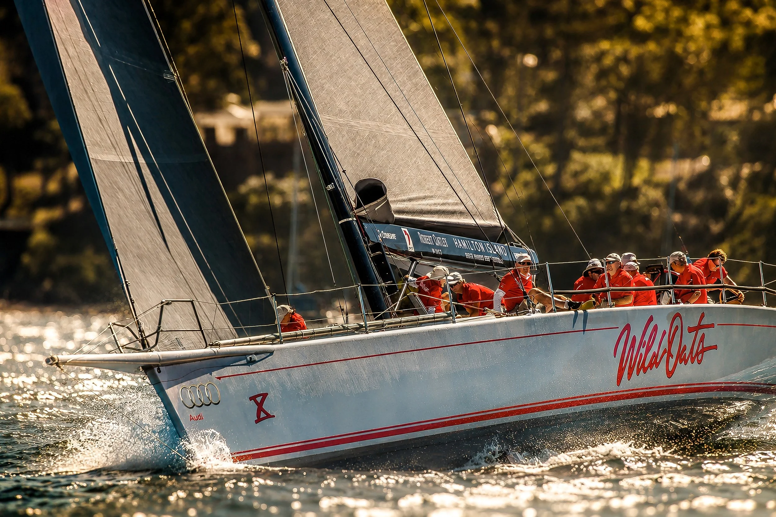A racing sailboat with a crew of sailors wearing red shirts and white hats, speeding through the water with large sails, during sunset or golden hour.
