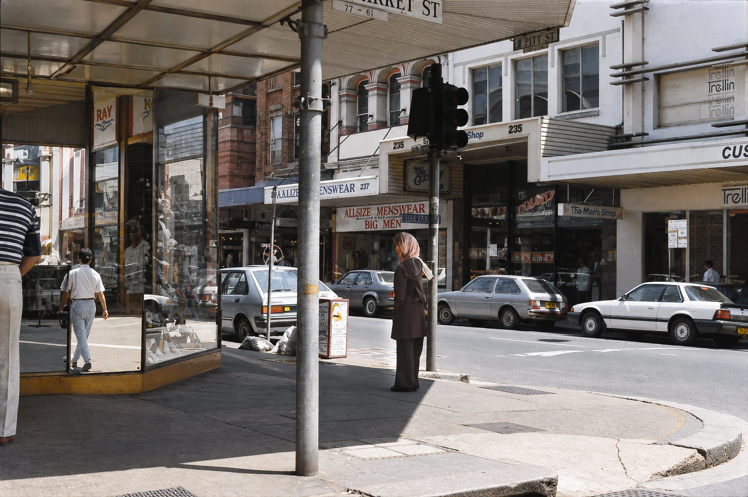 Sydney Shopping Street - A broad Sydney intersection reveals period cars, shopfronts, signs, pedestrians and the deep shadows of the covered pavement. The photograph documents the architecture and everyday commercial life of the city.
