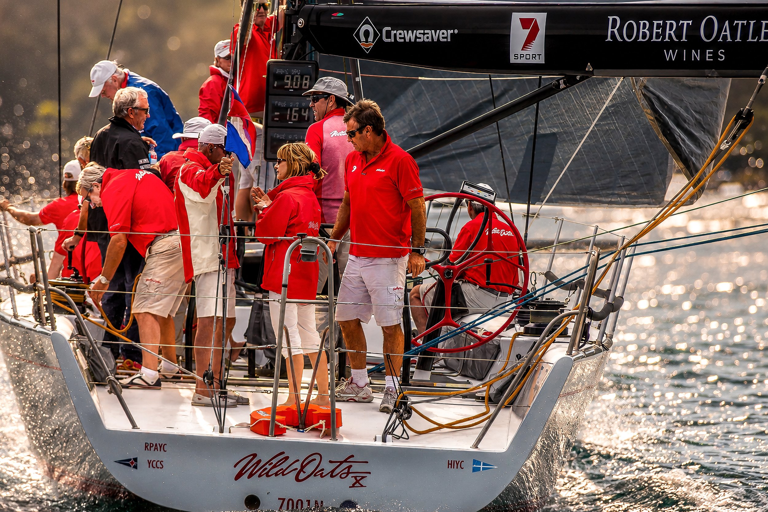A group of people on a sailboat racing on the water, wearing red and white clothing, some with hats and sunglasses, including a person holding a blue, white, and red flag. The sailboat has various brand logos and text, and the water sparkles in the s