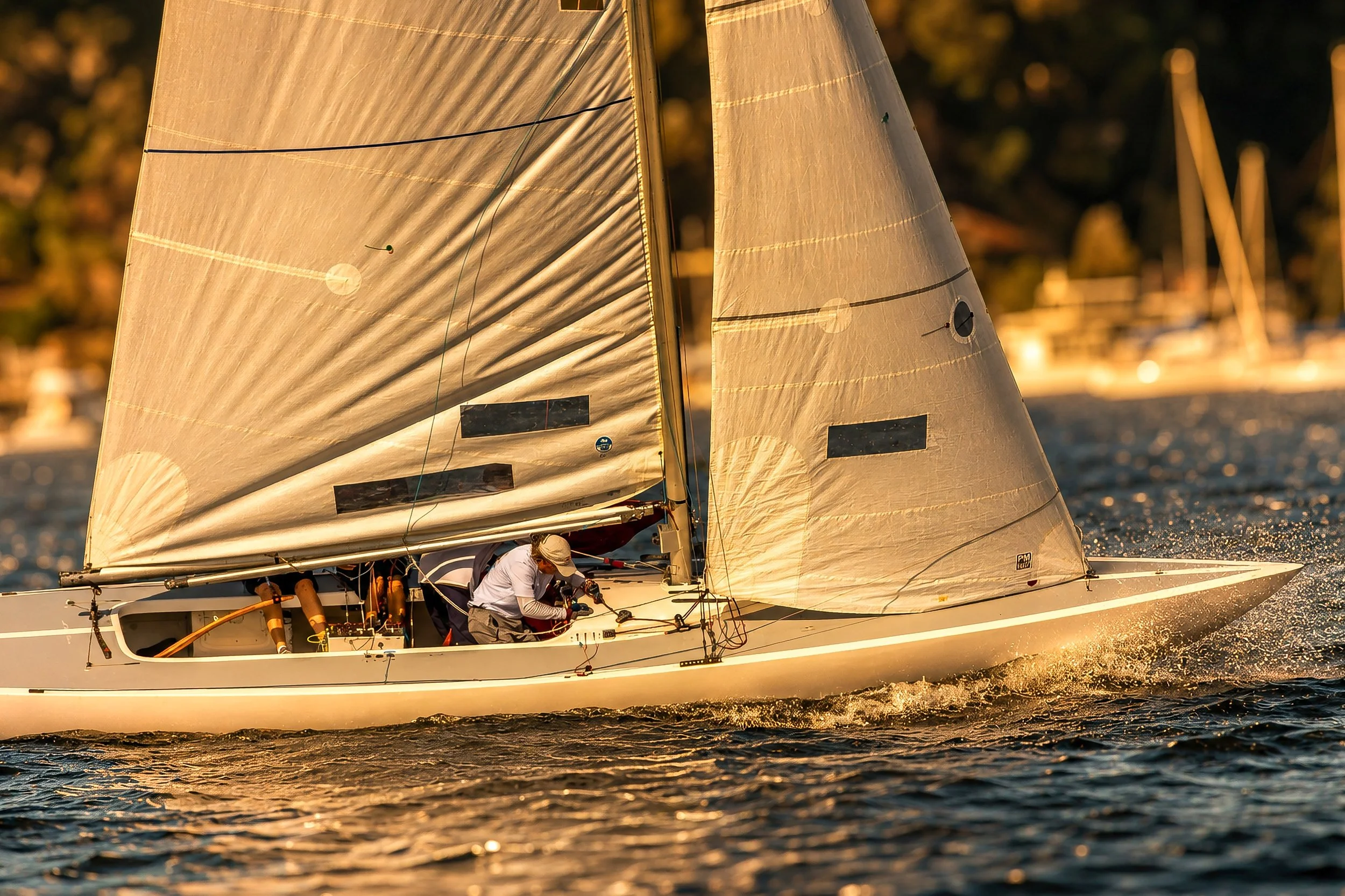 A sailboat with white sails racing on a body of water during golden hour sunlight, with a small crew onboard working on the boat as it cuts through the water.