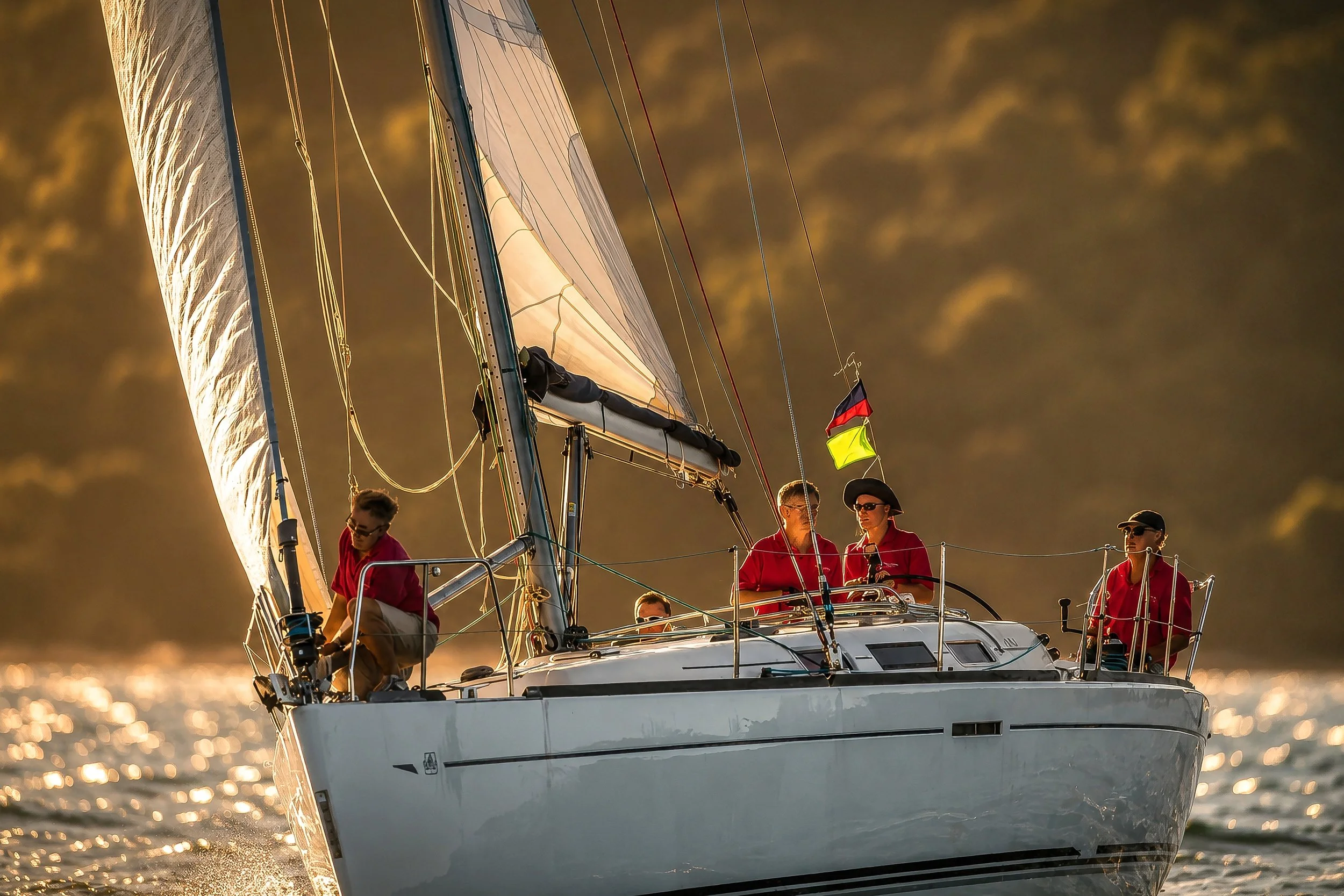 A sailboat with multiple people on board sailing on water during sunset, with the sunlight reflecting off the water.