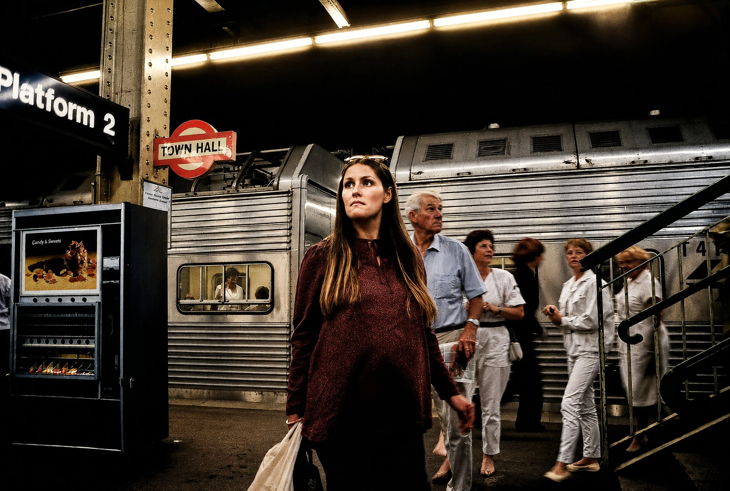 Women and men on a subway platform in New York City, with a sign indicating 'Platform 2' and a round 'Town Hall' station sign, along with a vending machine and stairs.