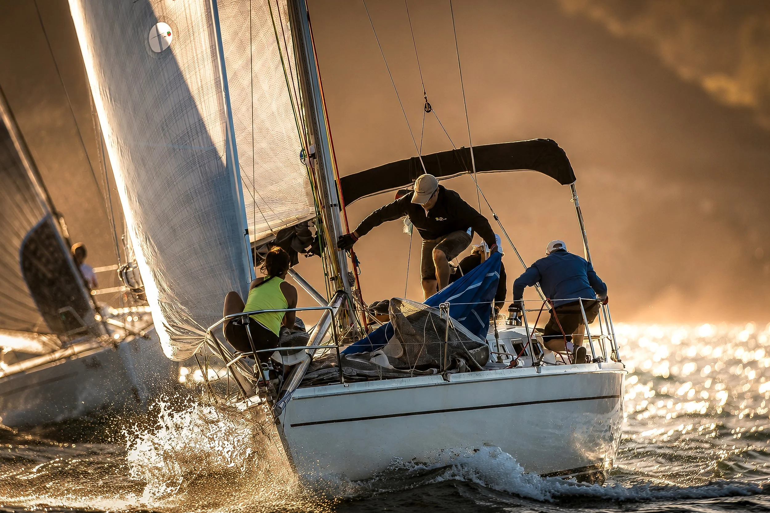 A sailboat with multiple people on board sailing on the water during sunset, with a close-up of the boat's hull and sails capturing sunlight.