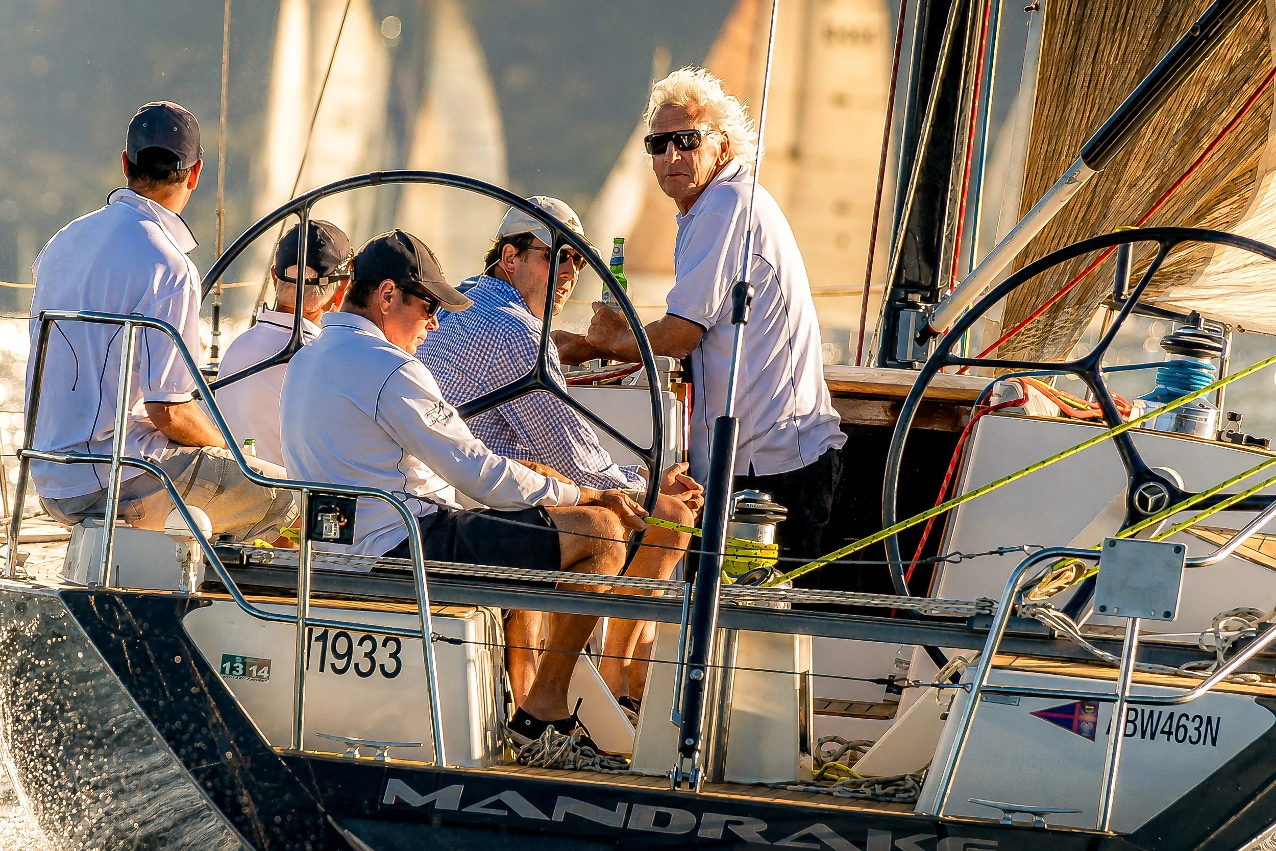 People sailing on a boat during sunset, with a man with white hair wearing sunglasses and a white shirt at the front, surrounded by other crew members on the deck.