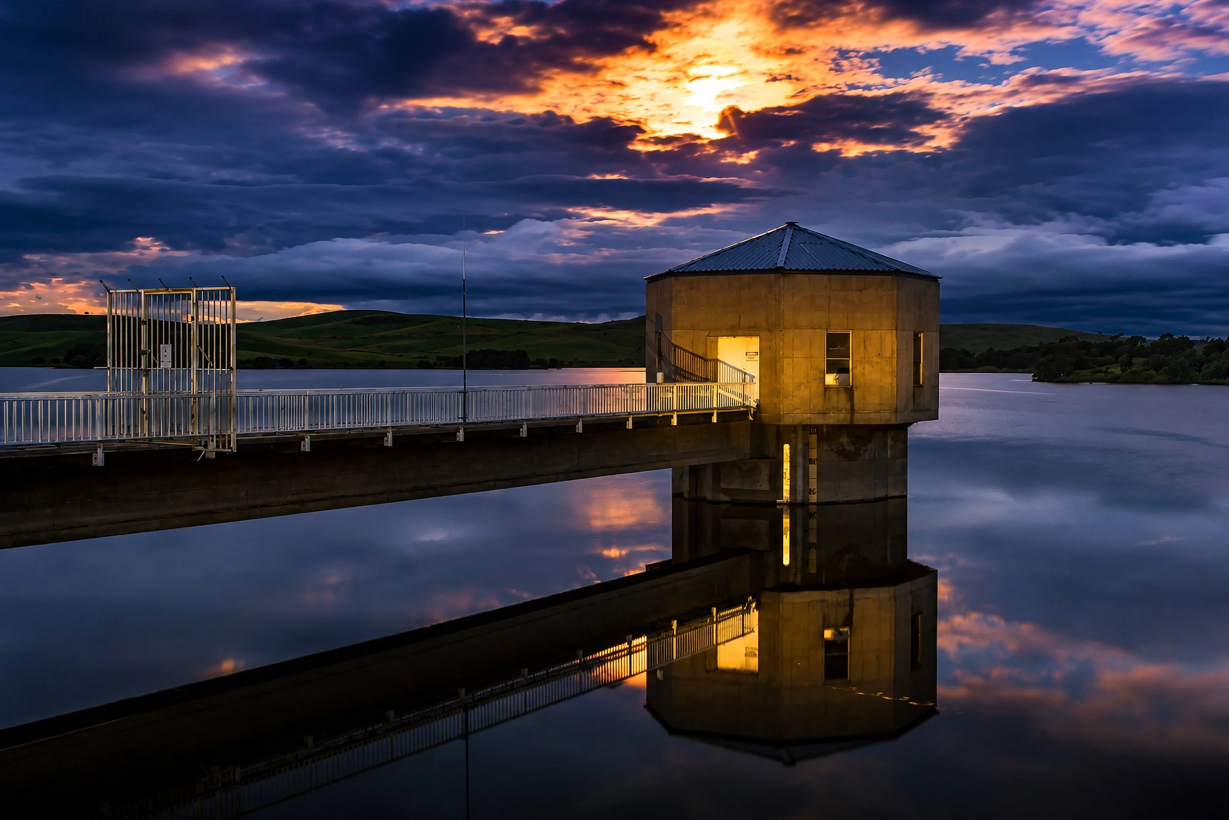 A tranquil pump house and jetty reflected in still water beneath a colourful twilight sky.
