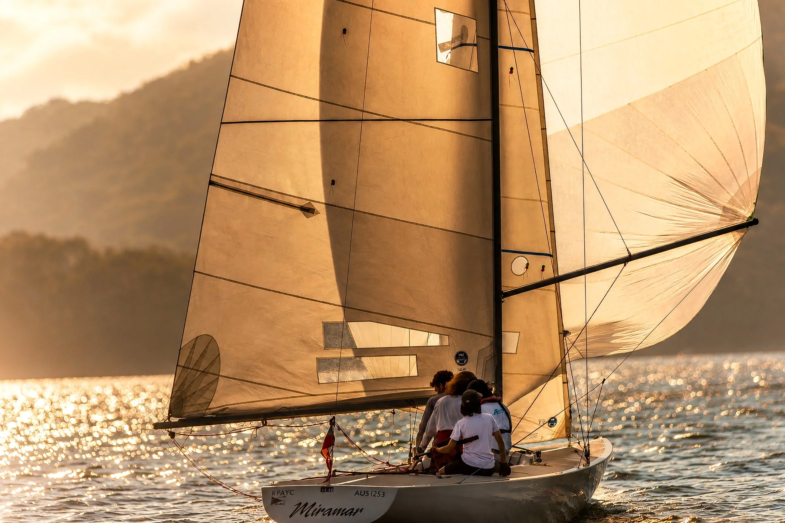 Sailboat with large beige spinnaker sails on shimmering water during sunset, with a crew of people on deck.