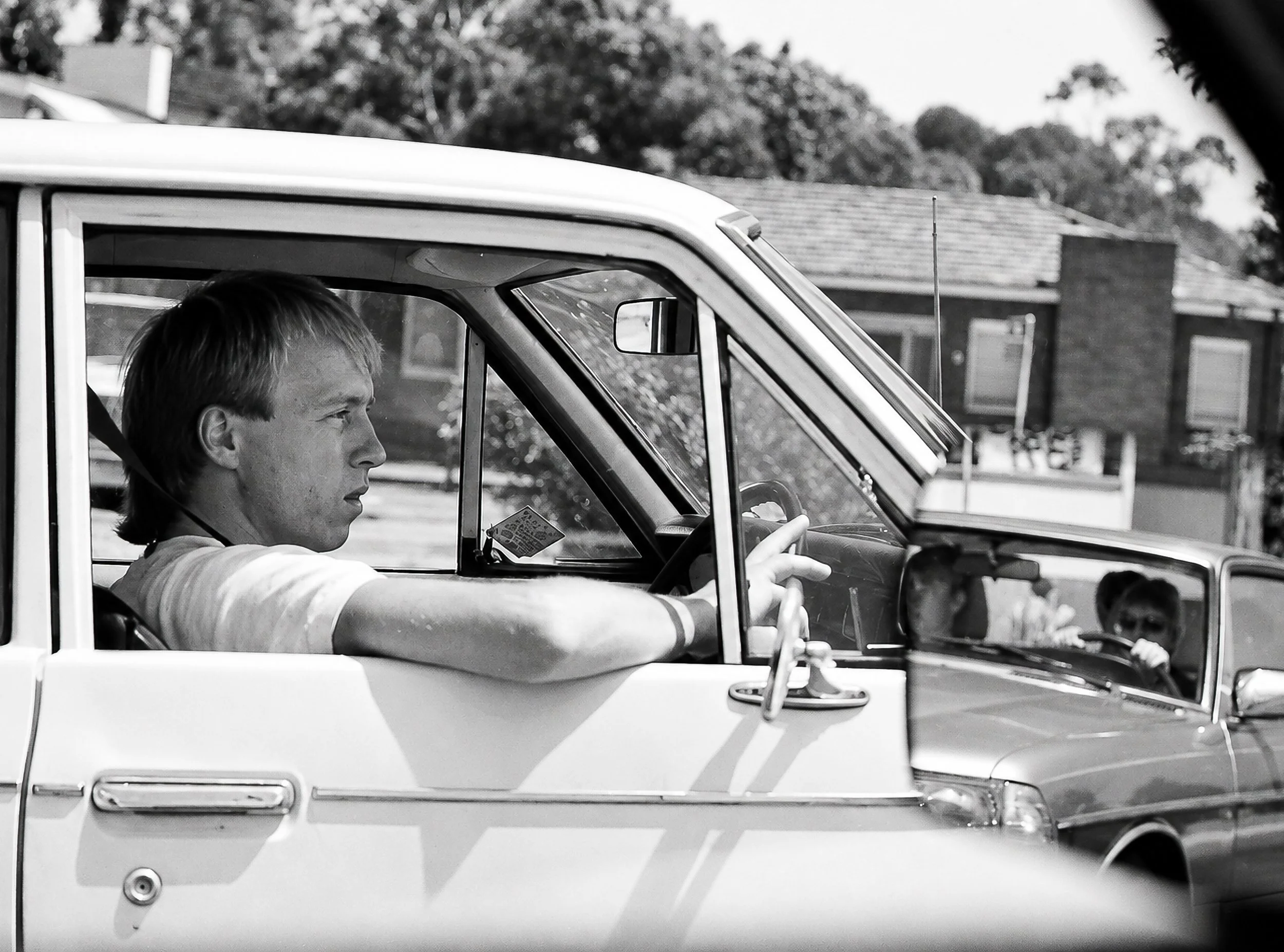 Waiting in Traffic - A driver sits at the wheel while another vehicle appears beyond him. Photographed from close range, the scene captures the ordinary rhythm of Sydney traffic and the fleeting opportunities to photograph strangers while waiting at 