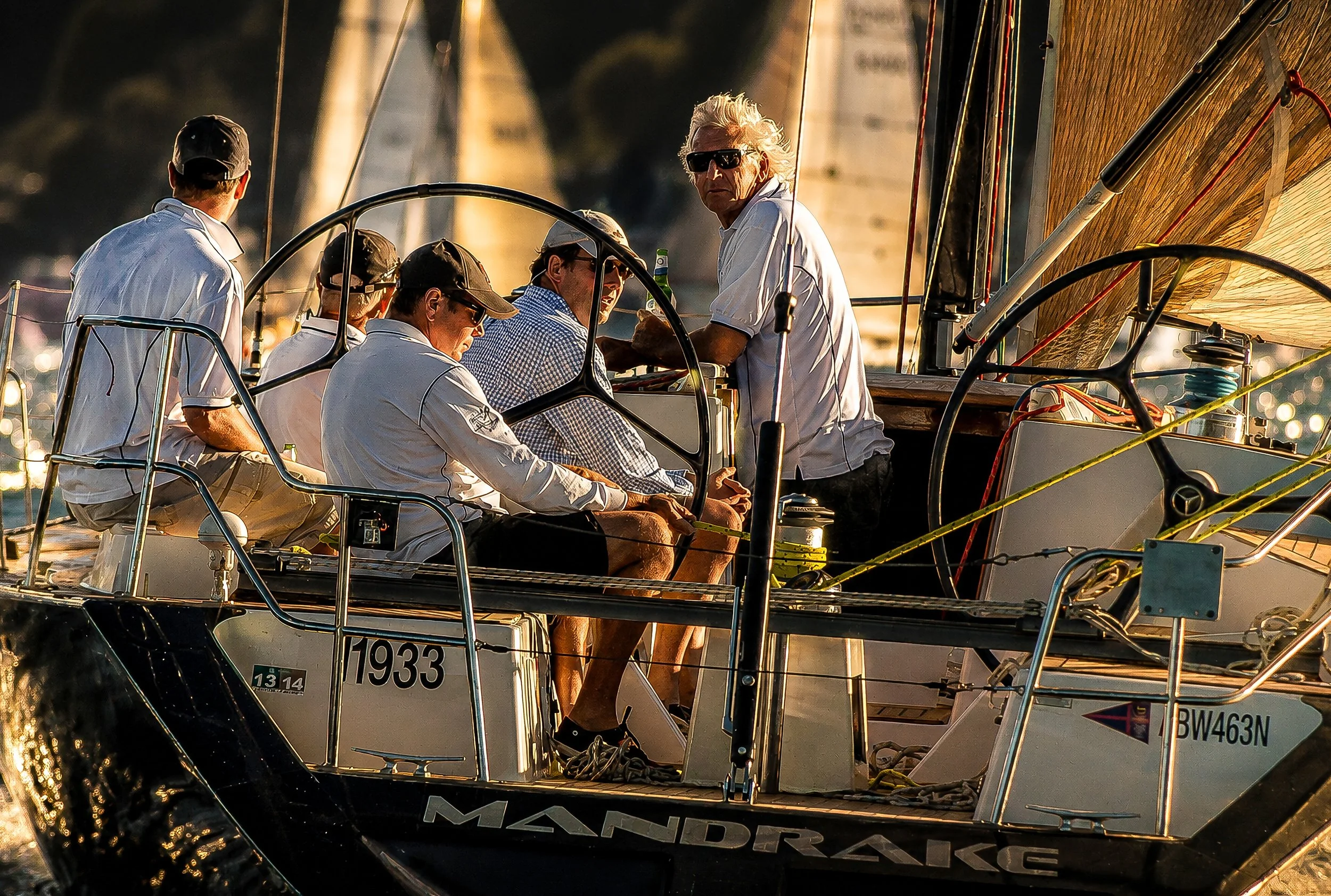 A group of five people on a sailboat, with one man with white hair and dark sunglasses standing and holding a bottle, and others sitting or standing around on the boat, with sails and rigging visible, during golden hour sunlight.