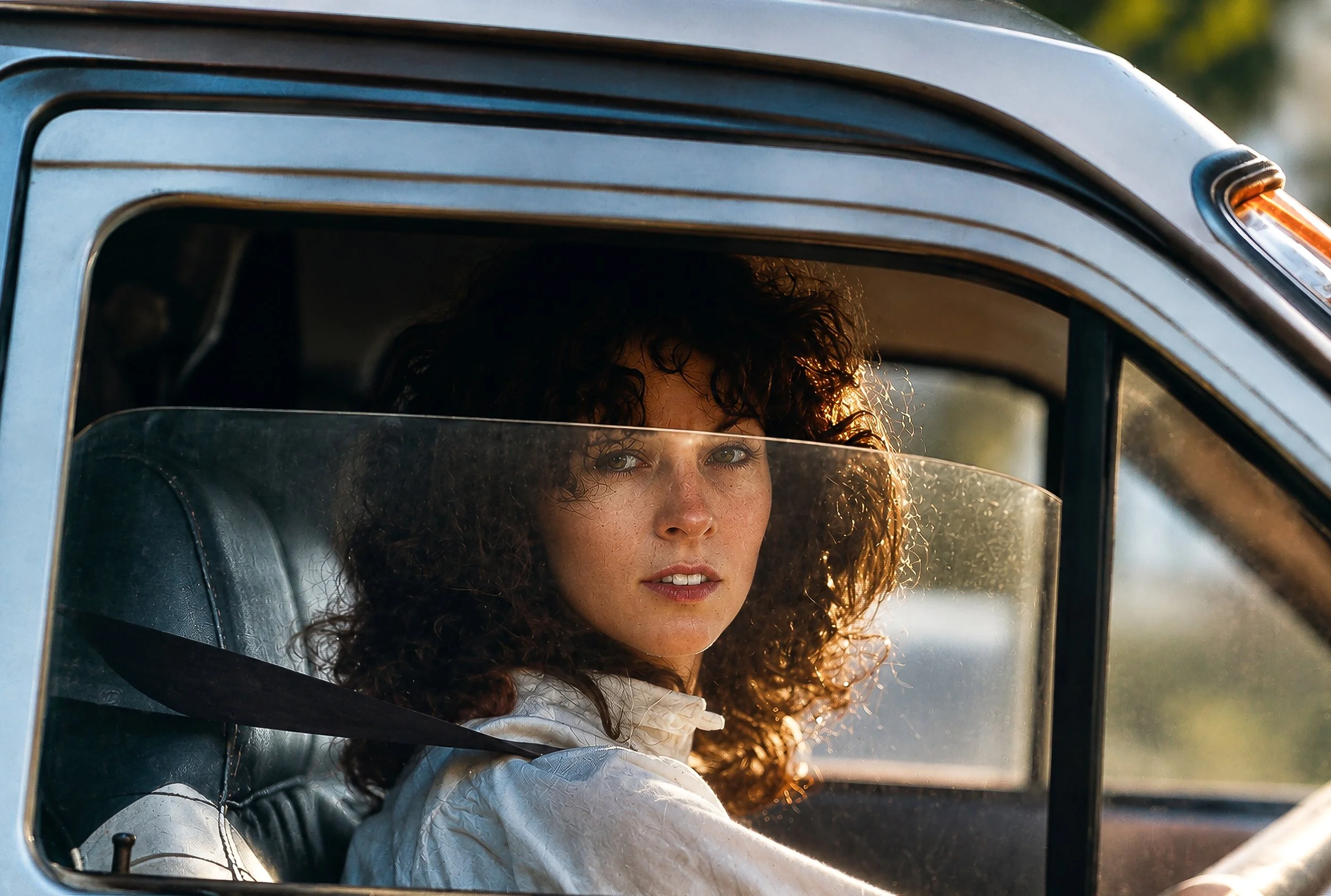 A woman with curly hair and light skin sits in the driver's seat of a blue vehicle, looking out of the open window with sunlight illuminating her face.