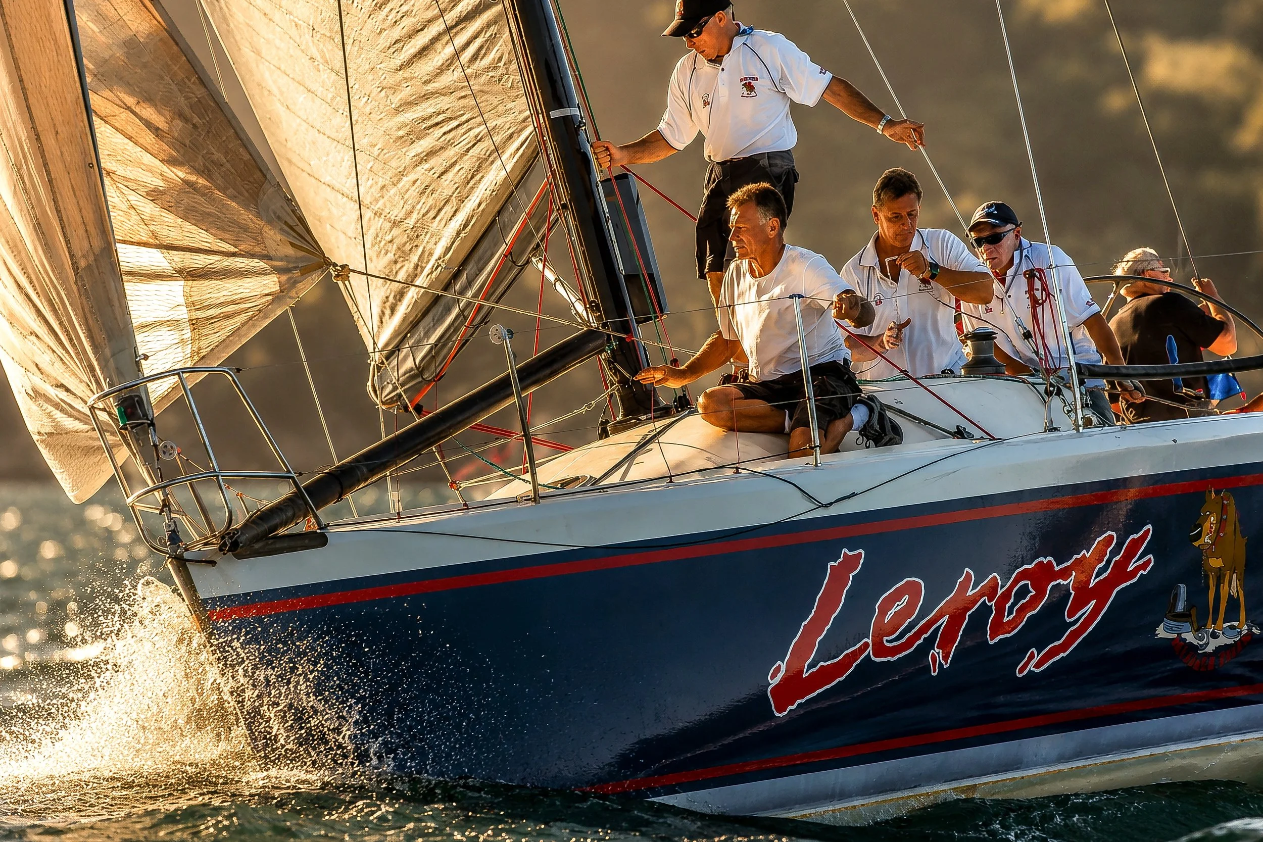 A sailboat racing on the water with a crew of five people managing the sails and steering, during sunset.