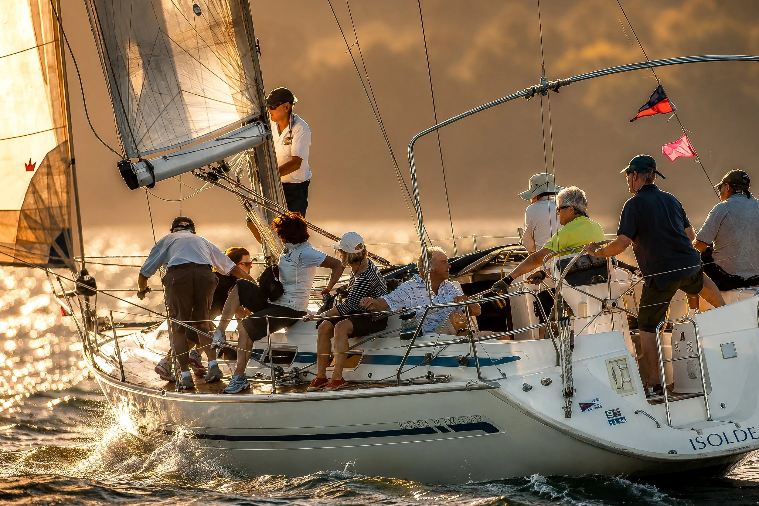 A sailboat with a group of people aboard, sailing on the water during sunset, with the sunlight reflecting off the water and the boat's sails.