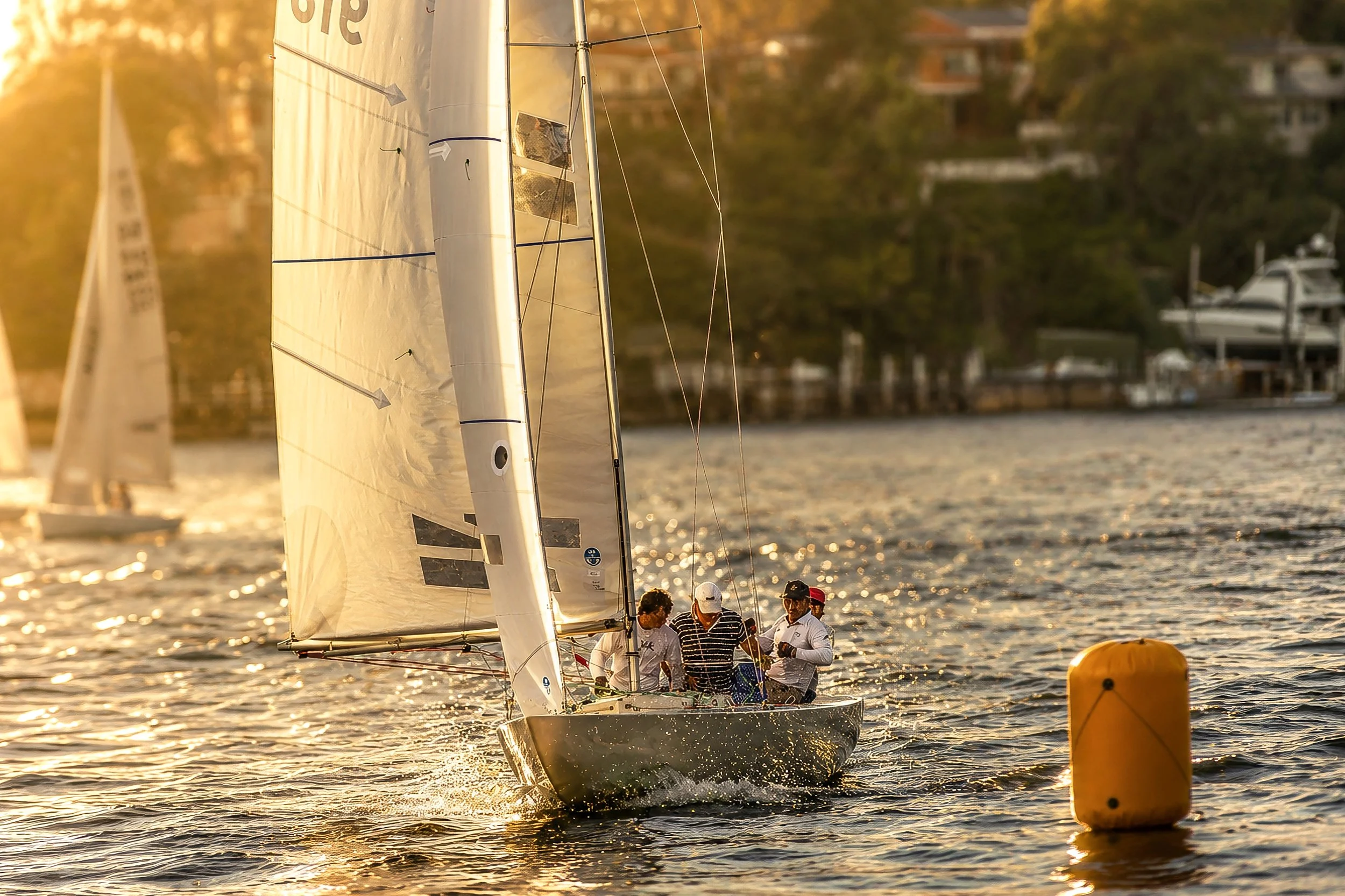 Sailing boat with a large sail and four crew members on a body of water during sunset, near a yellow buoy.