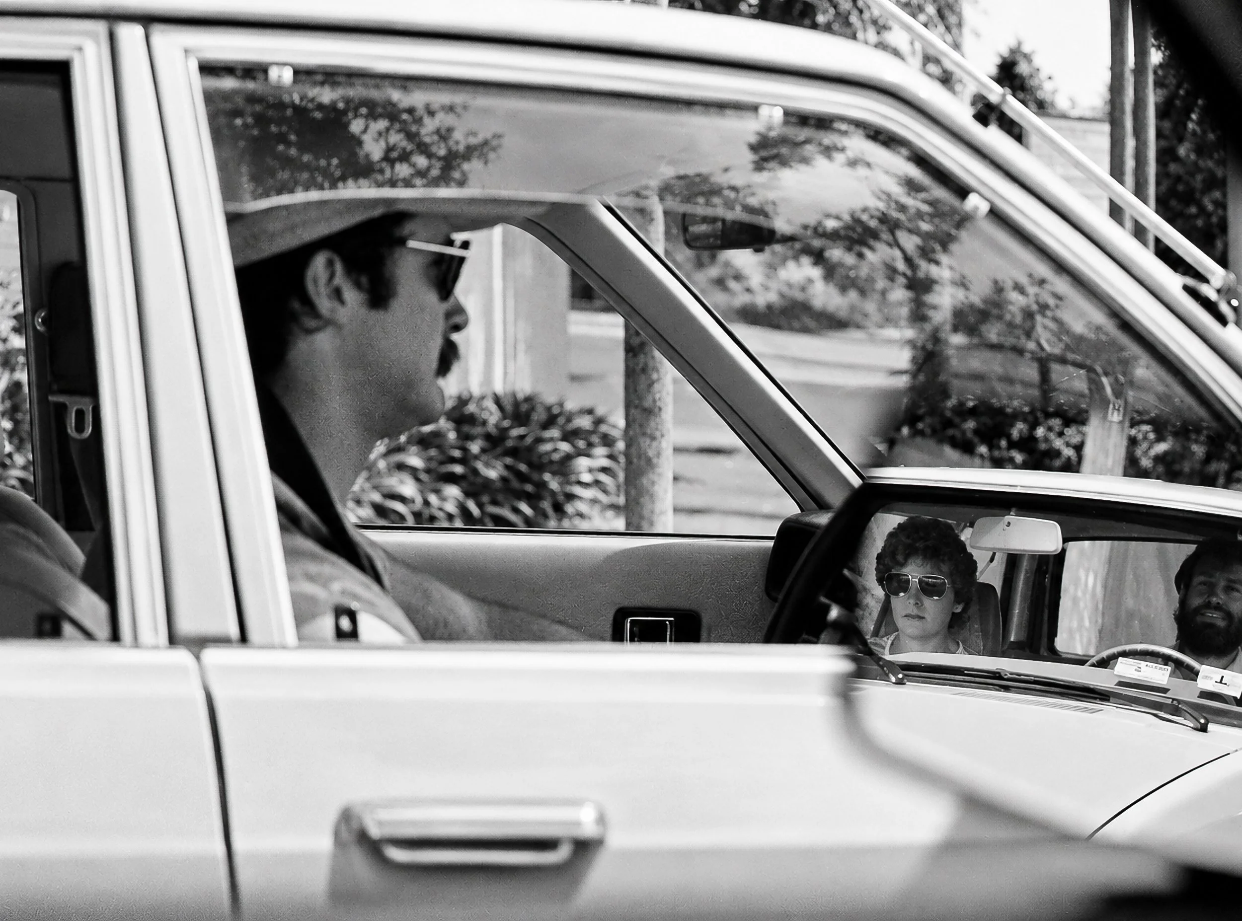 Two Cars, Two Encounters - A driver is photographed through the side of his car while another vehicle and its occupants appear in the wing mirror. The photograph combines several simultaneous moments of Sydney traffic within a single frame.
