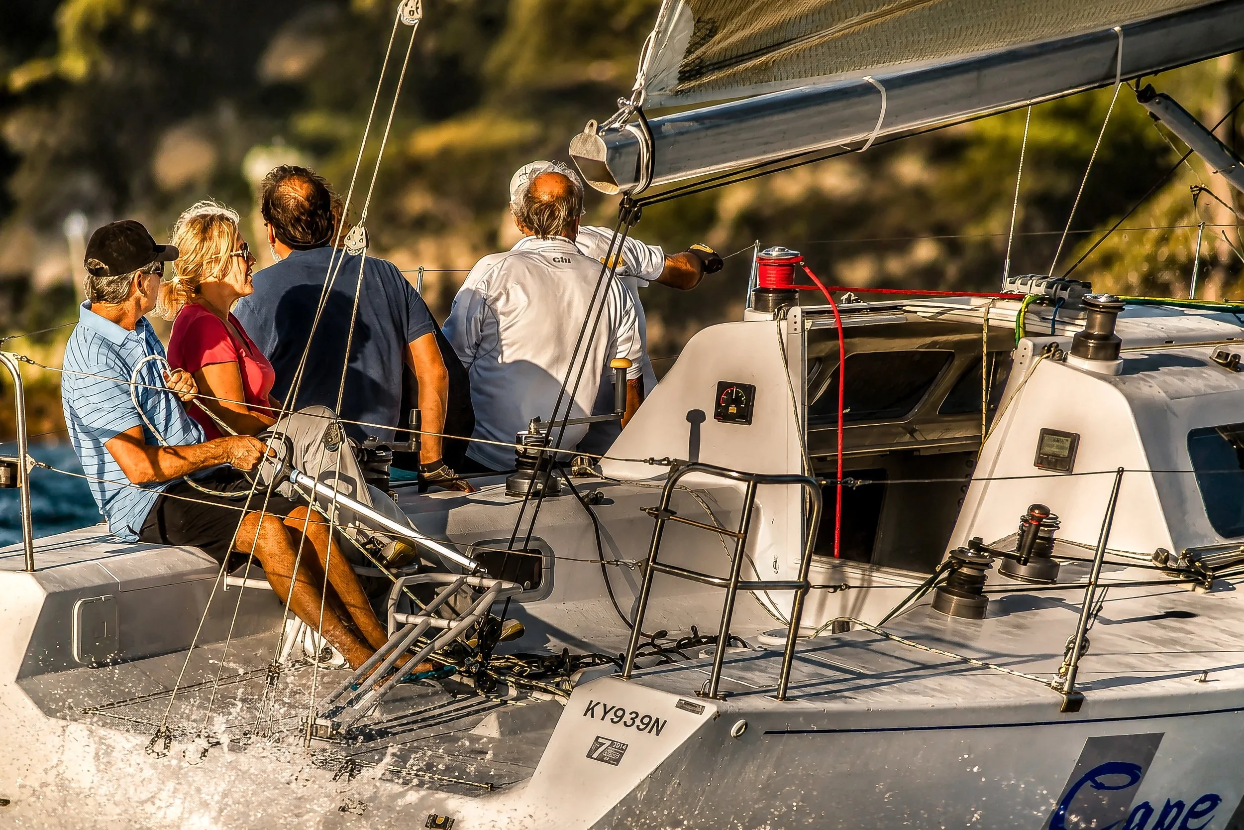 A group of people on a sailboat in motion, with water splashing at the bow. The boat has a white hull with the registration number KY939N. The crew is looking forward, with one person steering and others observing, as the boat navigates through water