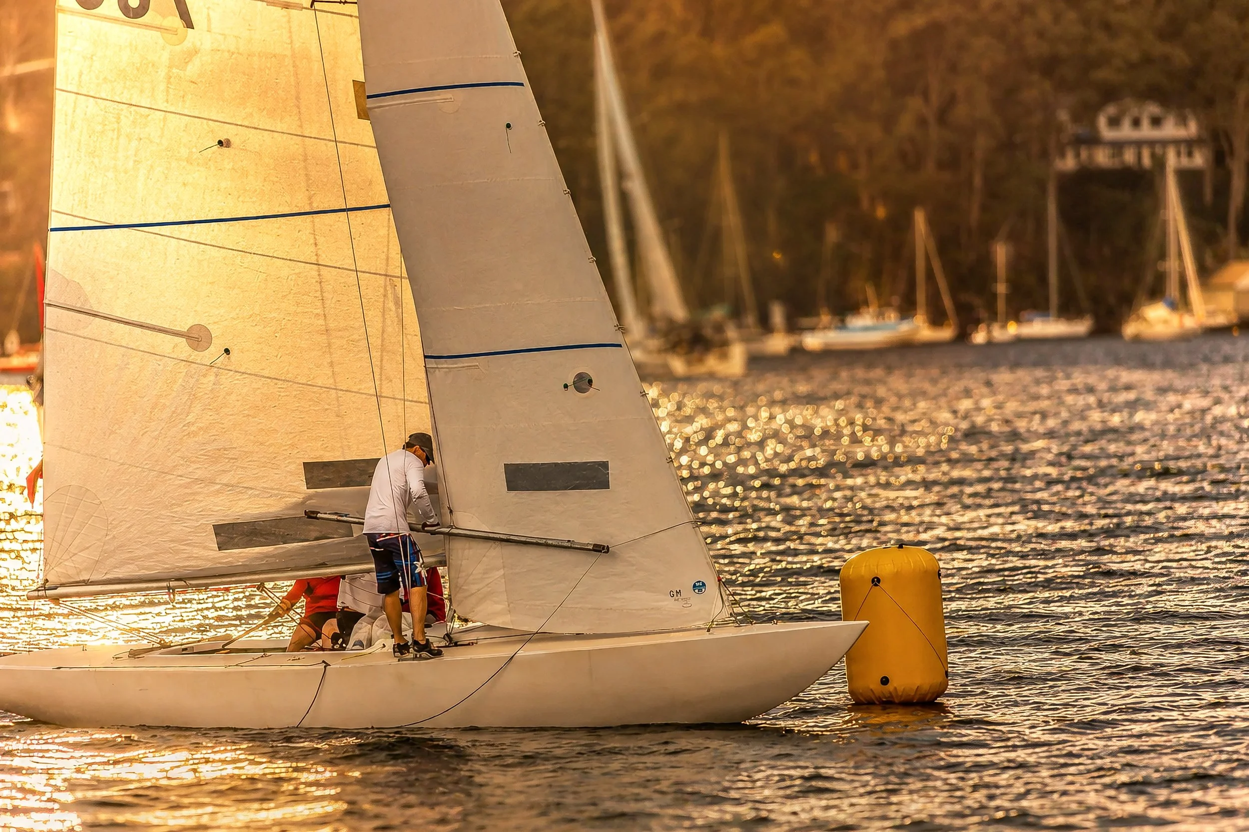 A sailboat with a white sail on shimmering water during sunset, with a person managing the boat and other sailboats in the background.