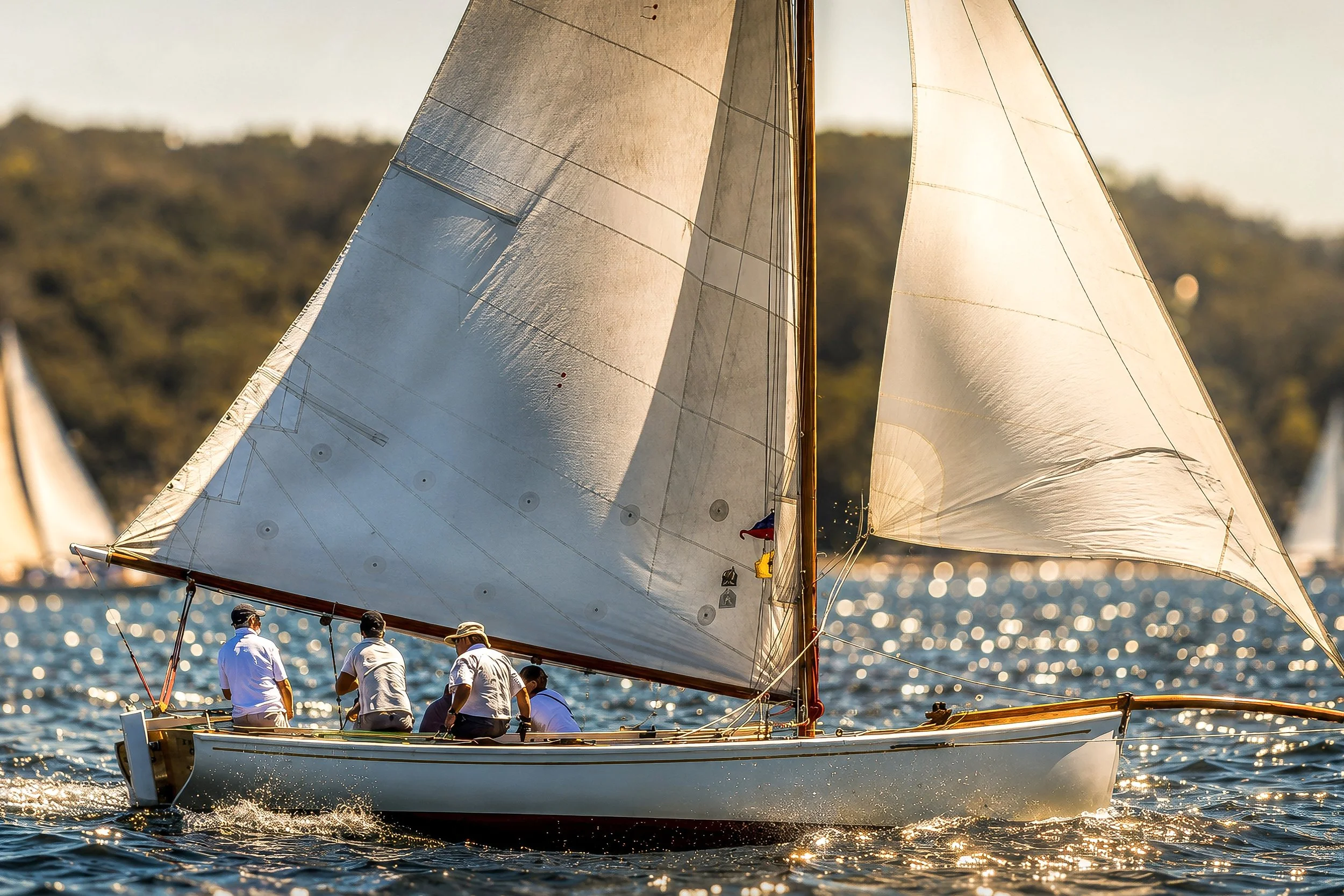 A sailboat with multiple crew members sailing on the water during sunset, with shimmering water and a distant shoreline in the background.
