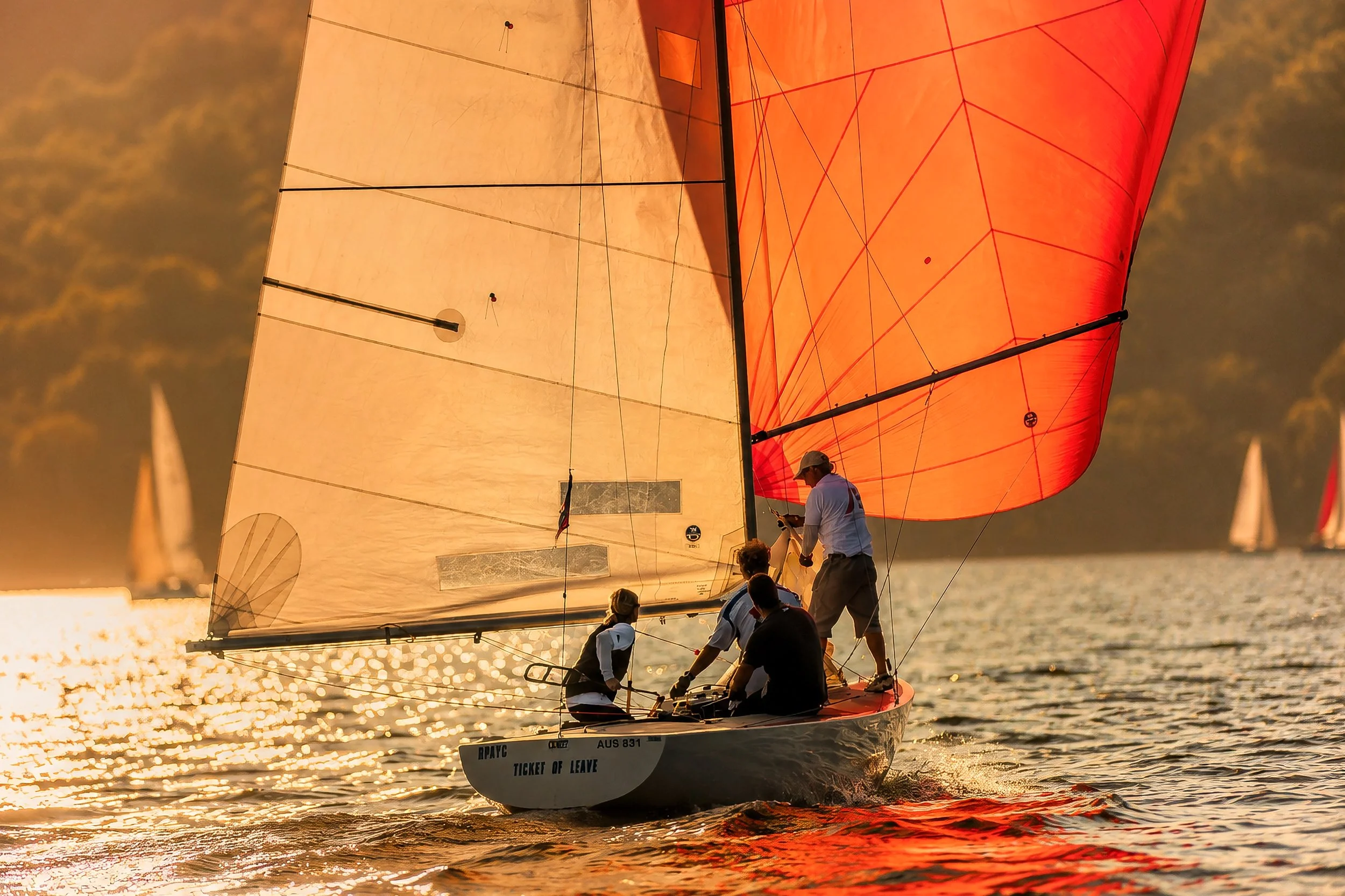 A sailboat with a red spinnaker sail and beige mainsail on the water during sunset, with people on board.