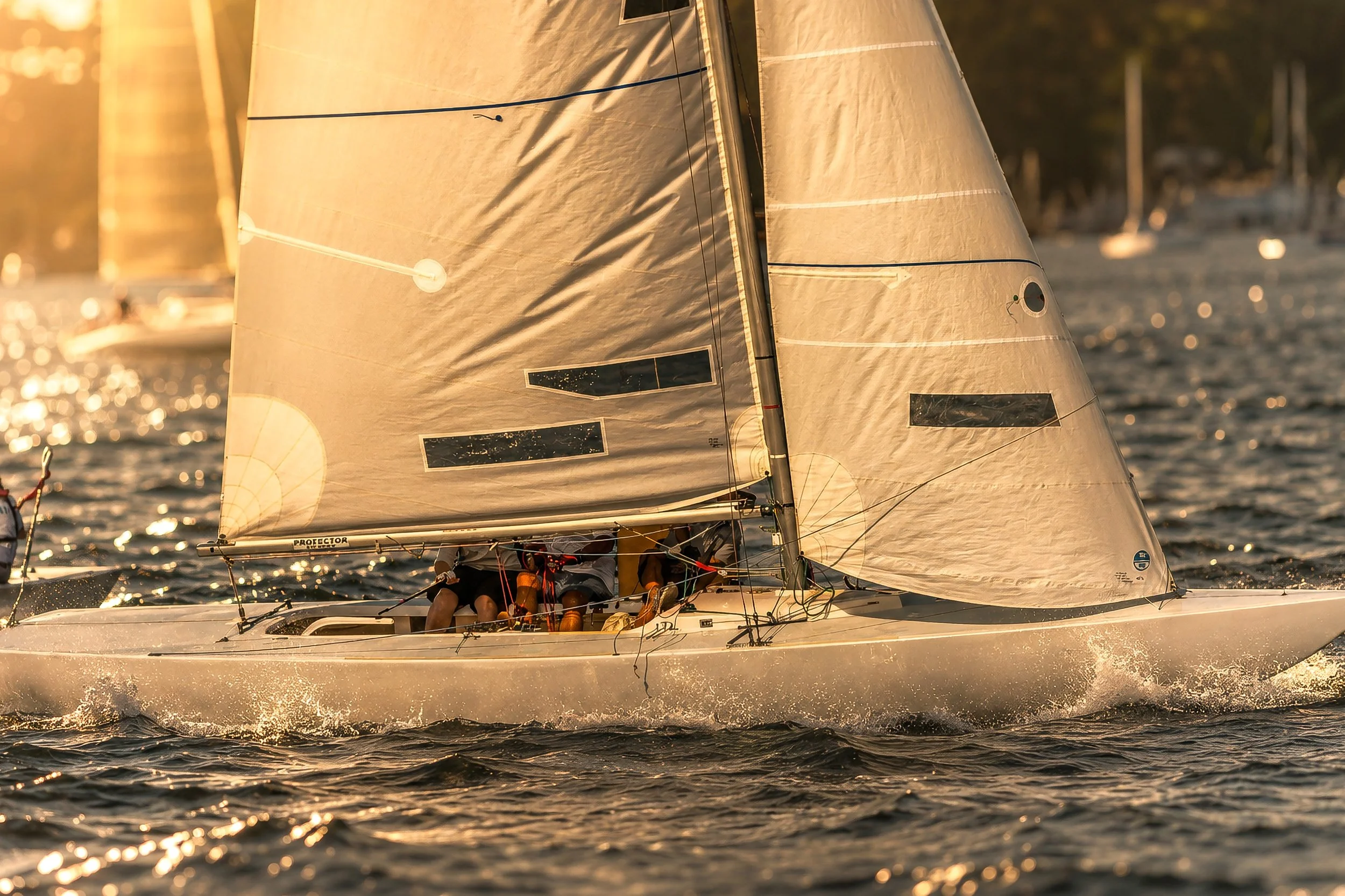 Sailboat racing on water during sunset with crew members on board, sails catching sunlight, scenic background of trees and boats.