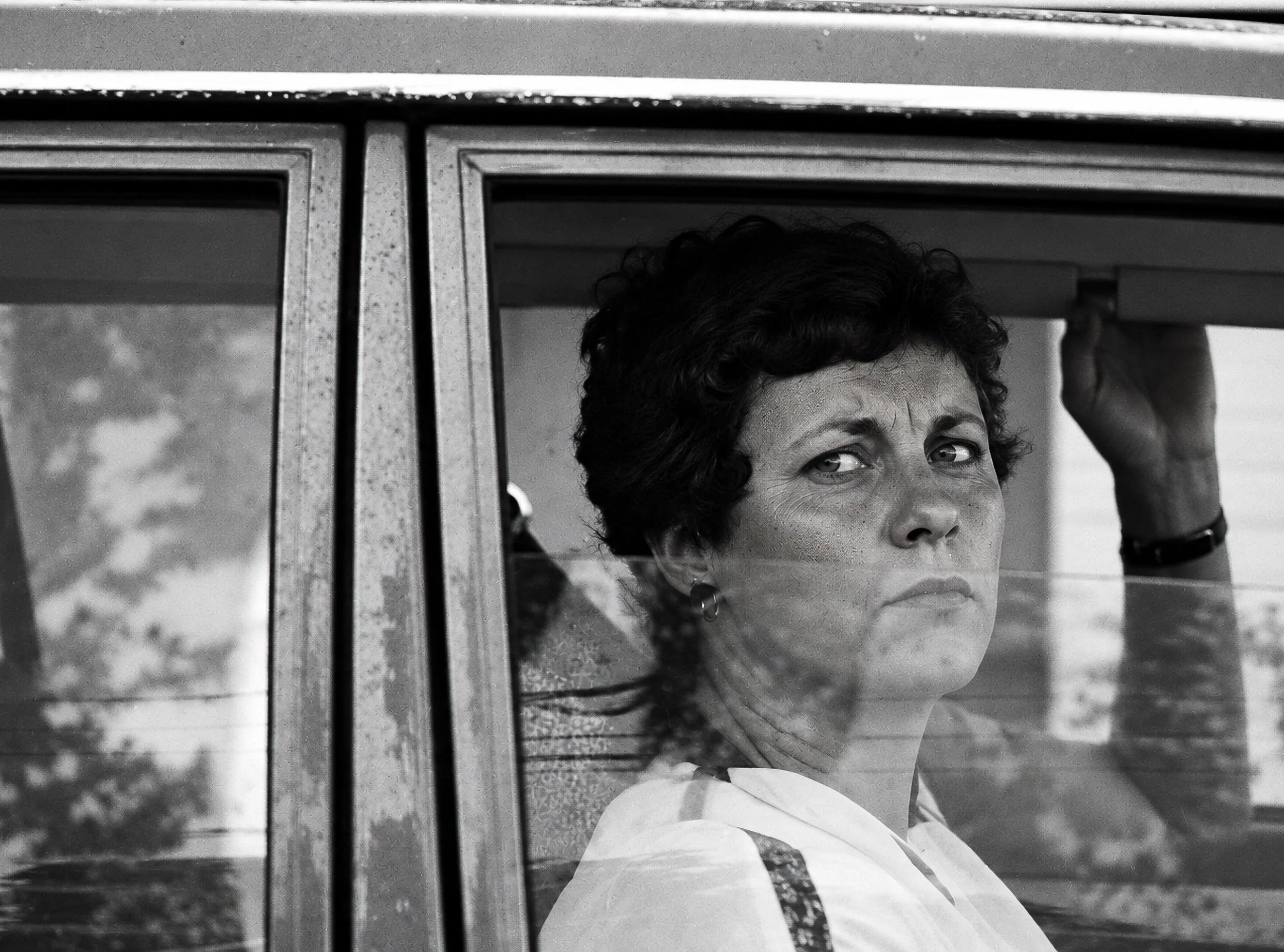 Behind the Glass - A woman looks directly towards the camera through a car window, with reflections of the surrounding street passing across her face. The glass separates photographer and subject while simultaneously becoming part of the photograph, 