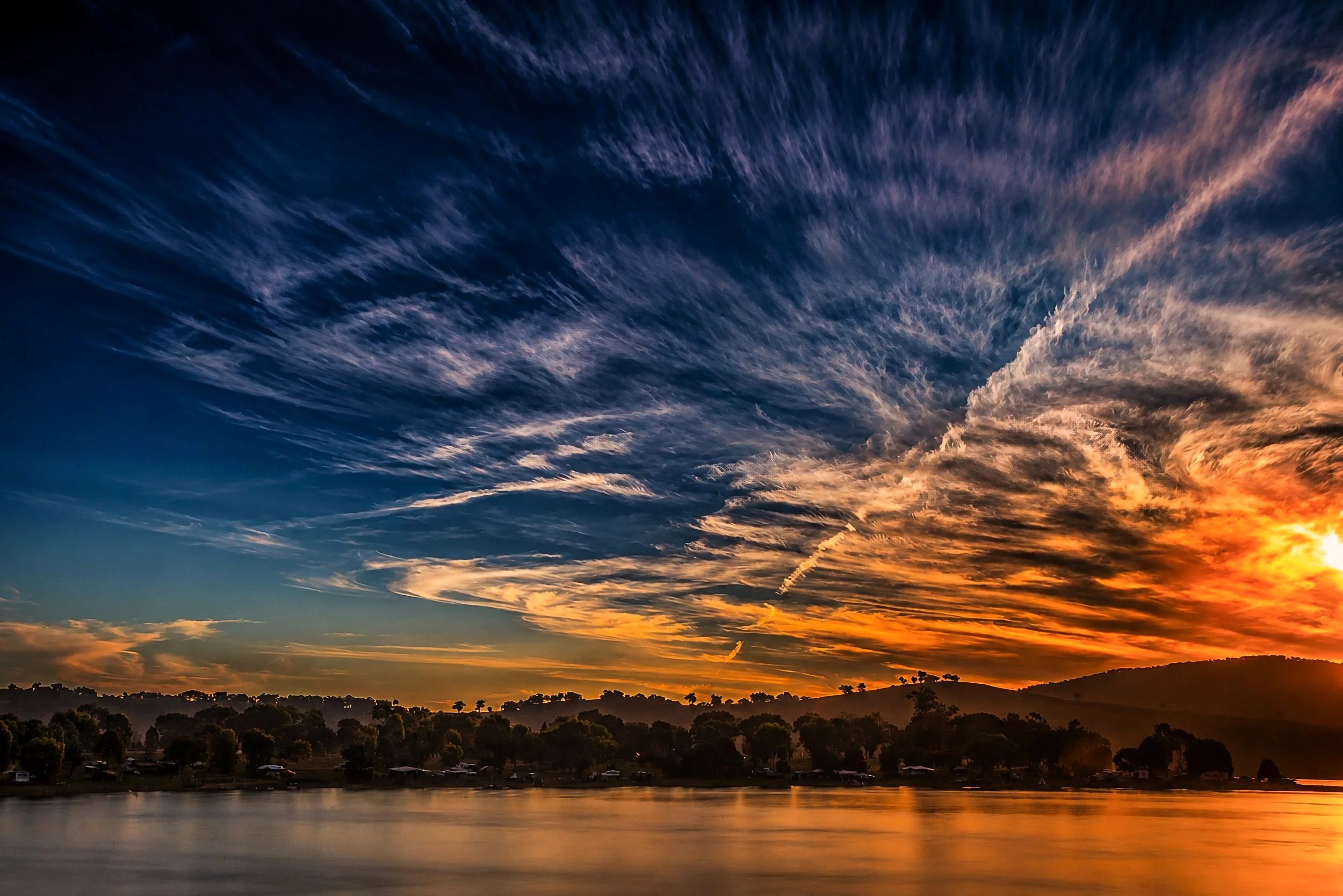 A spectacular Australian sunrise fills the sky with sweeping orange and blue cloud formations reflected across calm water.


