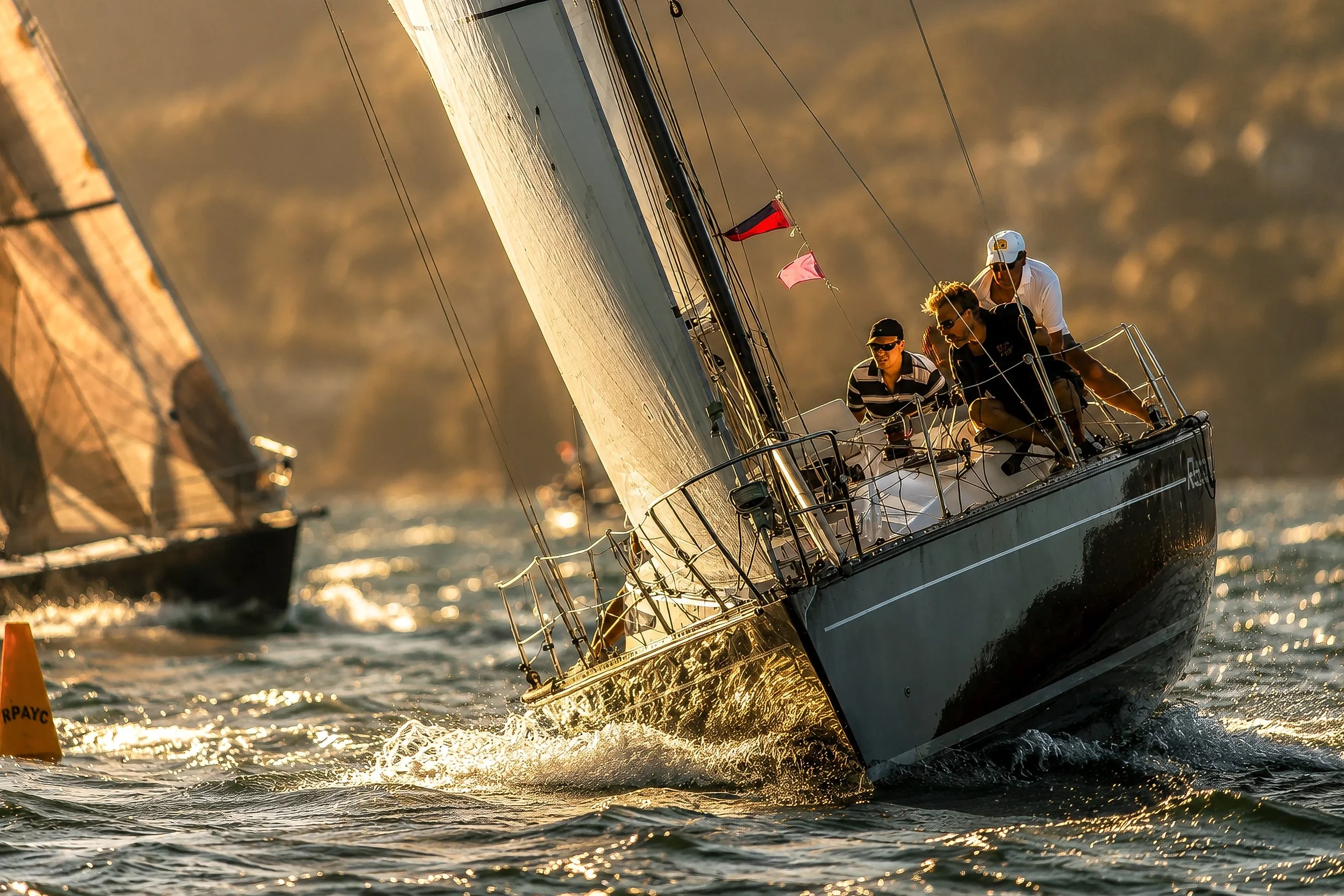 Sailing boat with crew members navigating through water during sunset or sunrise, with a blurred background of another boat and a marker buoy nearby.