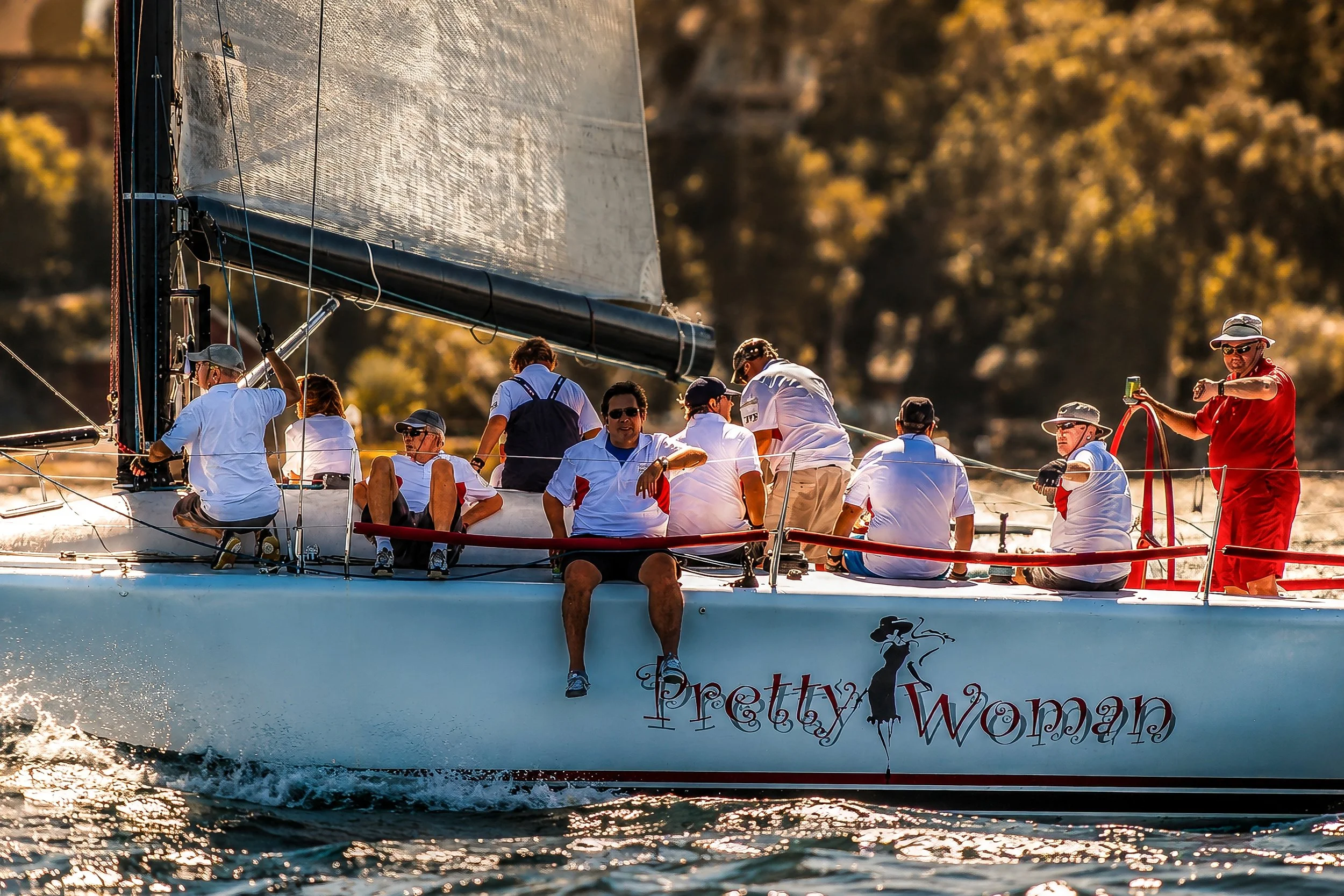 A group of people, mostly wearing white shirts, sailing on a boat during sunset. One person is taking a photo with a smartphone. The boat has a visible logo and a partly visible web address.