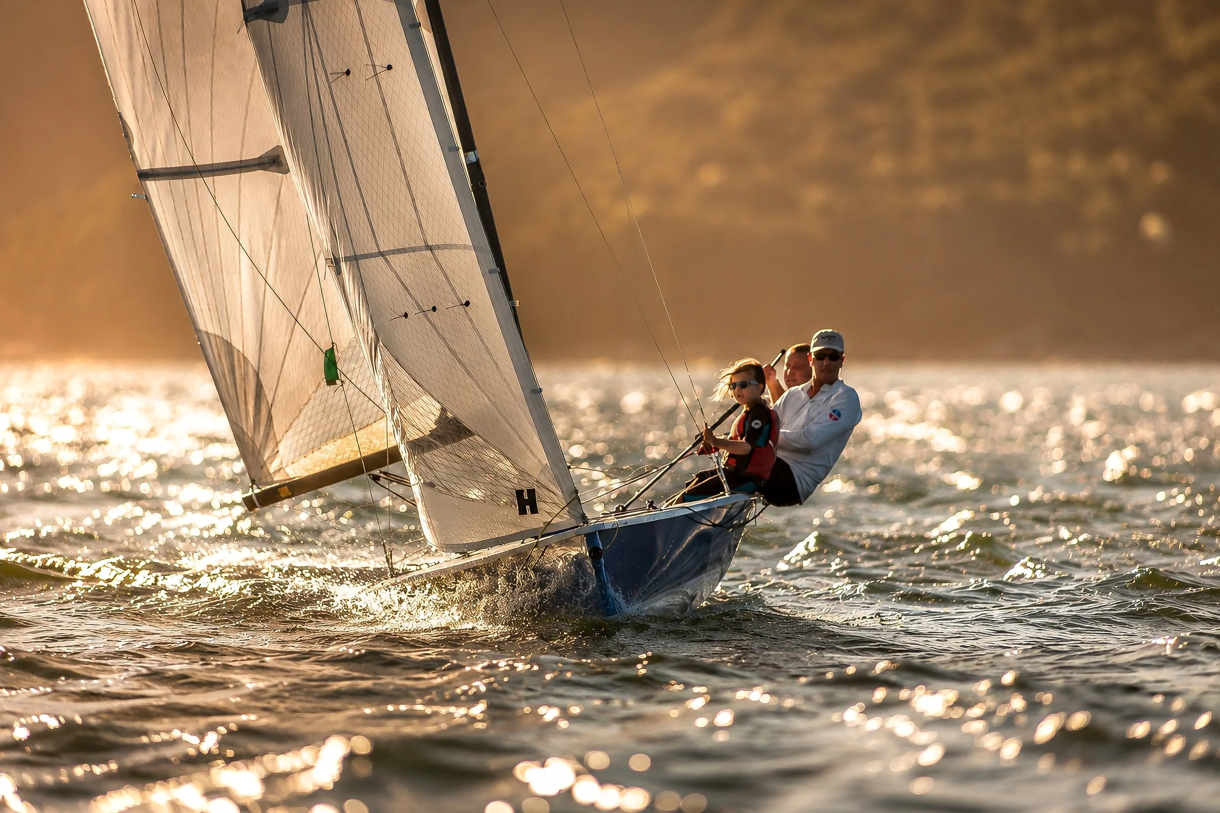 A small sailboat with two people, a man and a young girl, sailing on a body of water during sunset. The boat has large sails catching the wind, and the water sparkles in the sunlight.