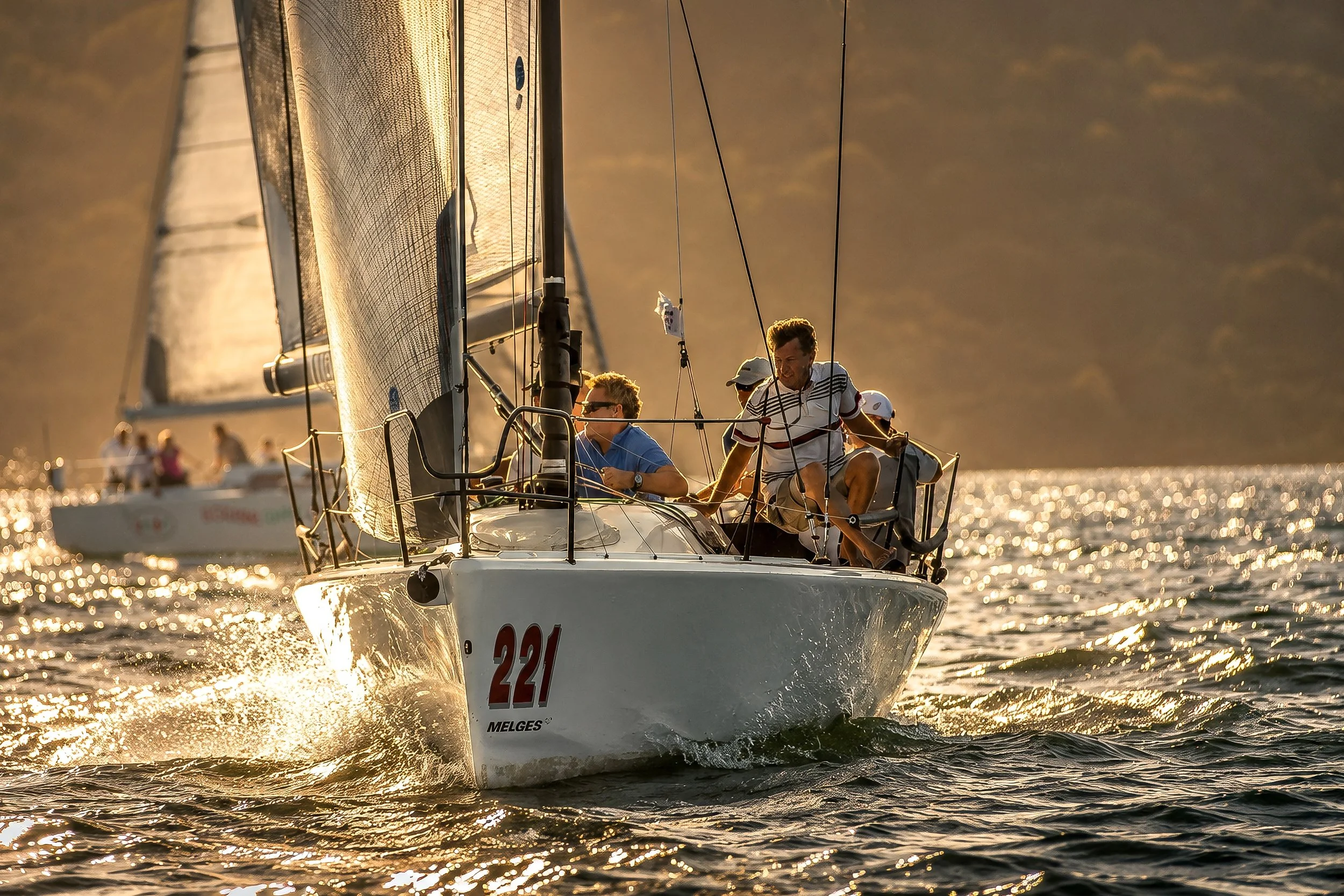 A sailboat with a white hull numbered 221, sailing on the water during sunset, with people on board and one person standing on the side.