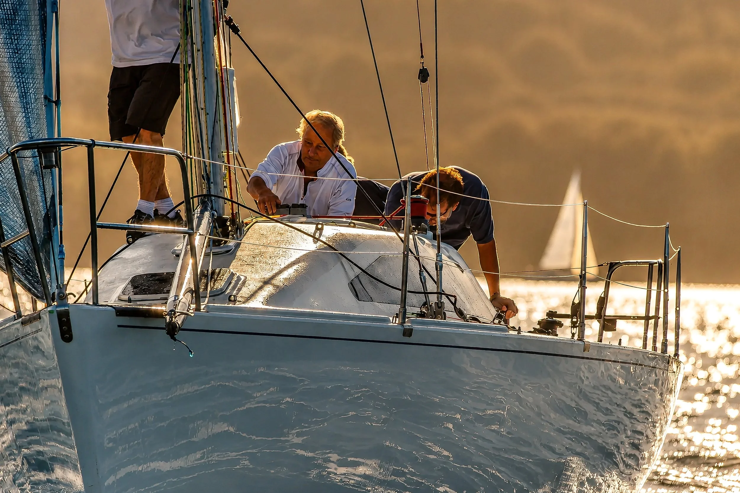 Two people onboard a sailboat with several ropes and equipment, sailing during sunset or sunrise on calm water.