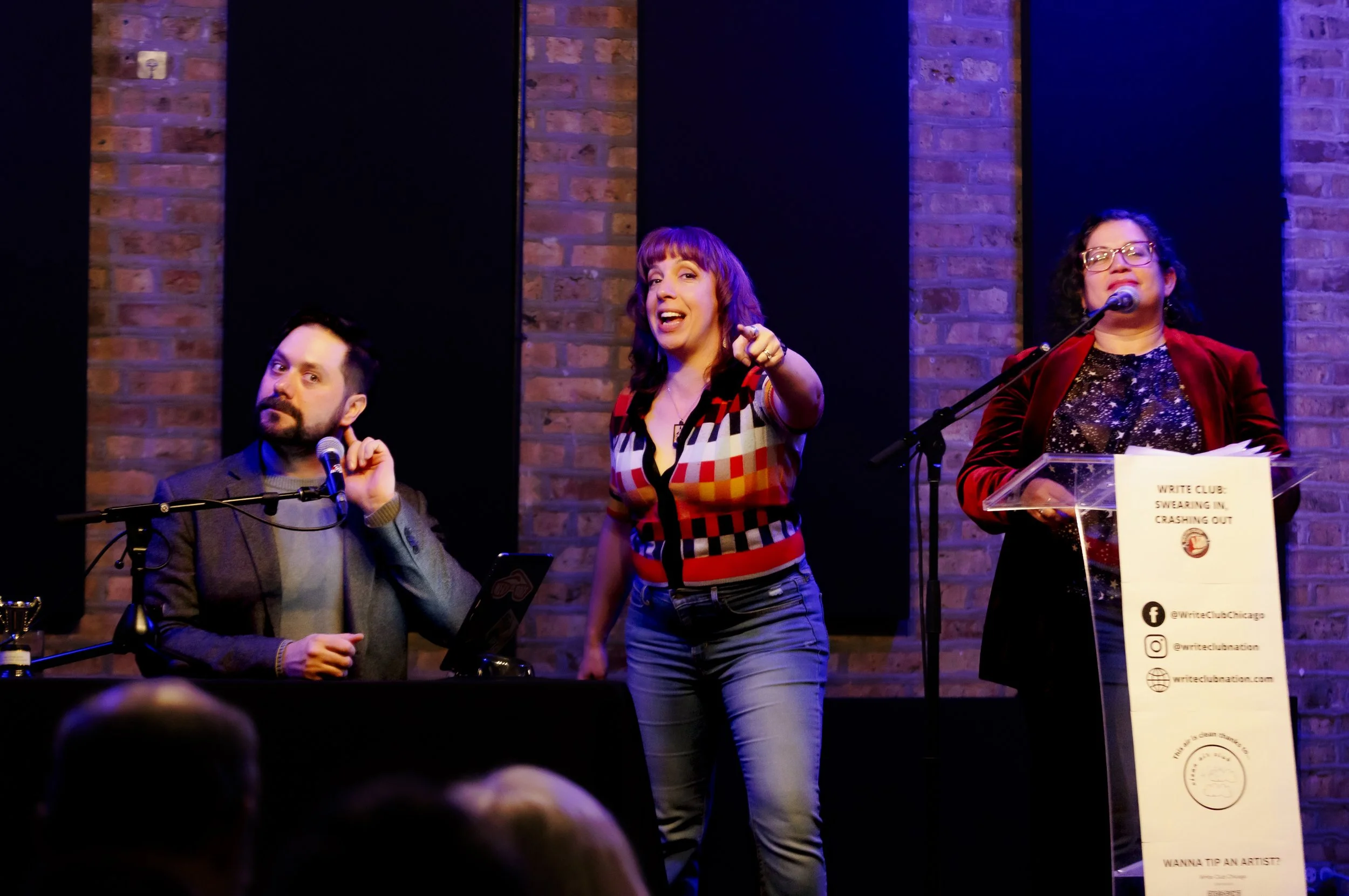 Far left, a timekeeper holds his finger to his ear to listen, center the host points to the audience, far right a combatant readies herself to perform a 7 minute essay