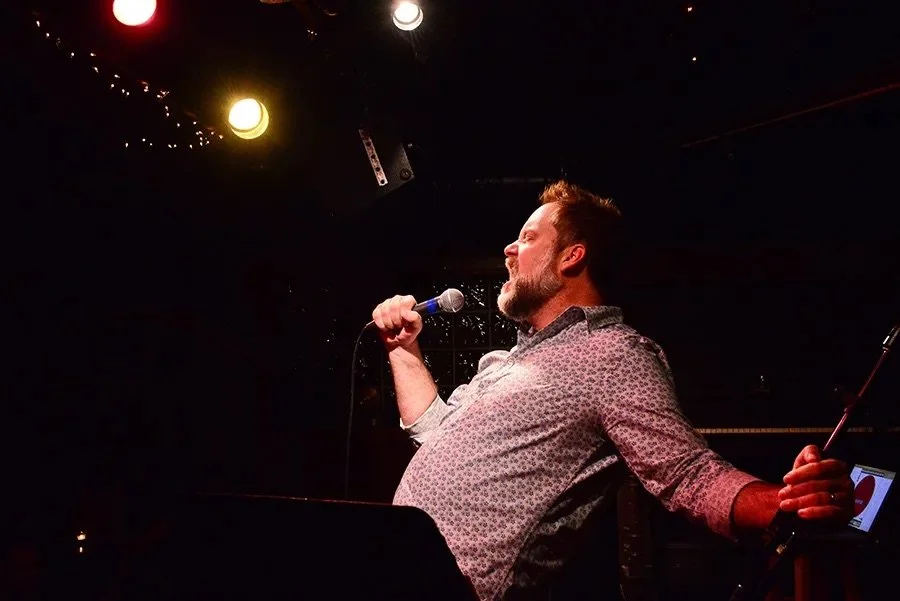 Write Club Founder Ian Belknap, a white male with a beard and mustache and wearing a patterned button down, leaning back and holding the mic, screaming for live lit