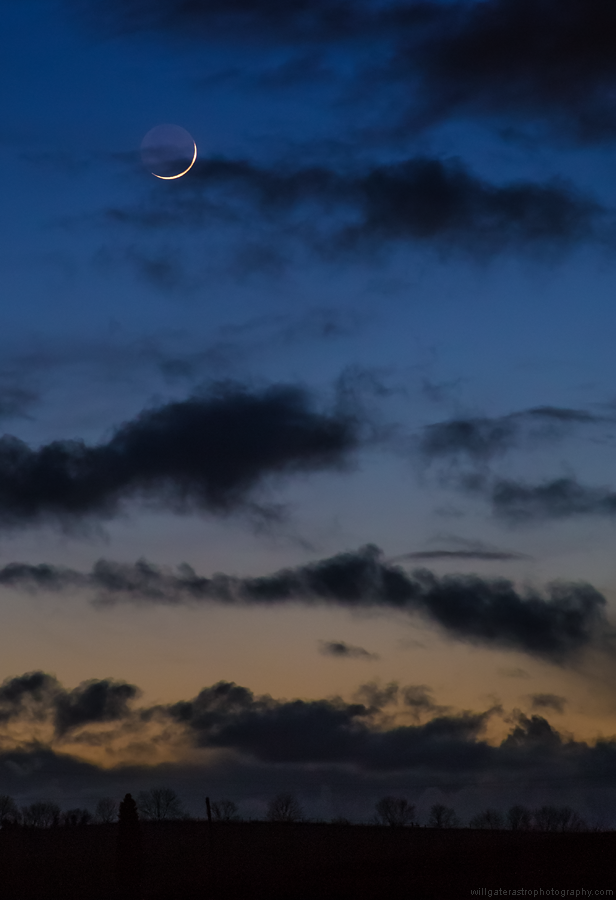 Twilight crescent Moon with Earthshine behind silhouetted clouds