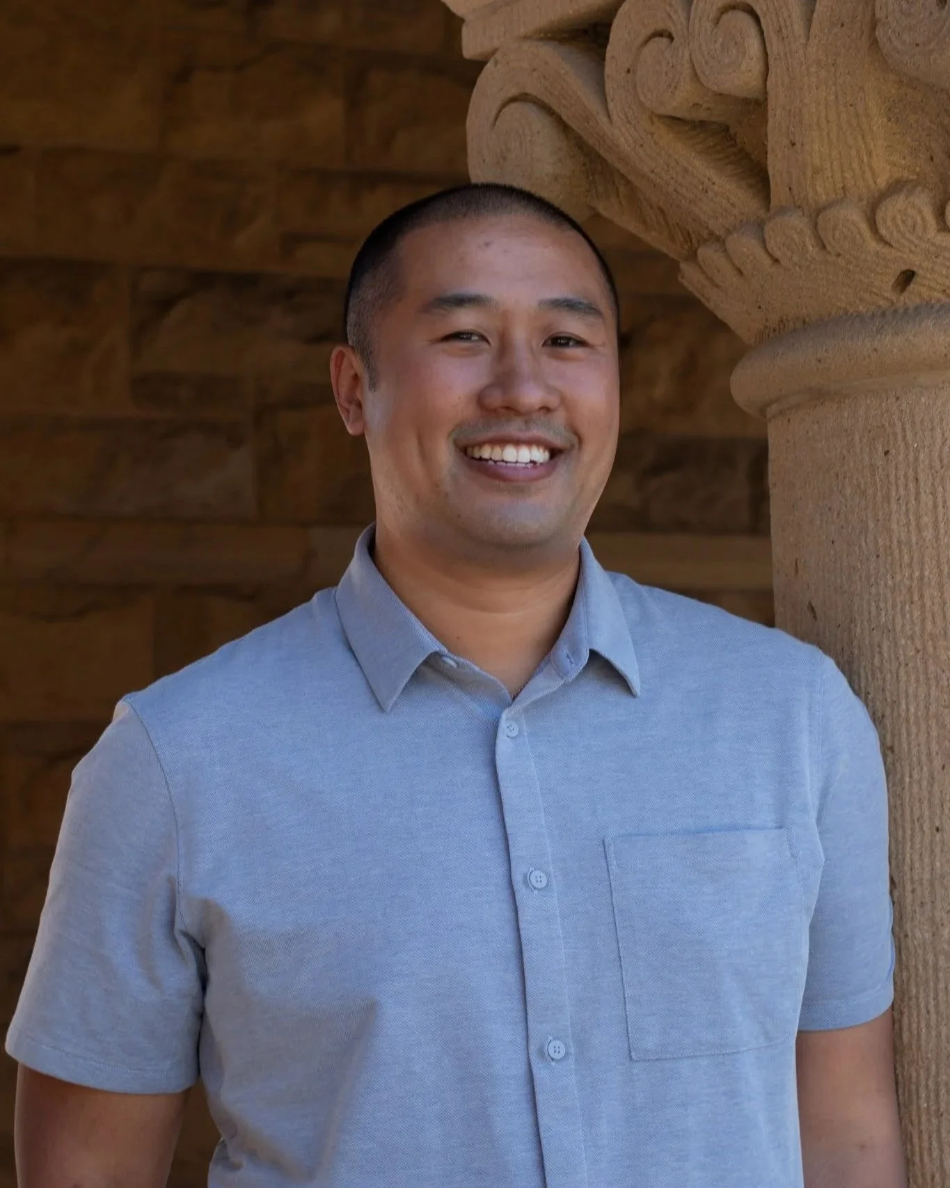 A smiling man with short hair wearing a light blue button-up shirt, standing next to a carved stone pillar with intricate designs, in front of a wooden background.