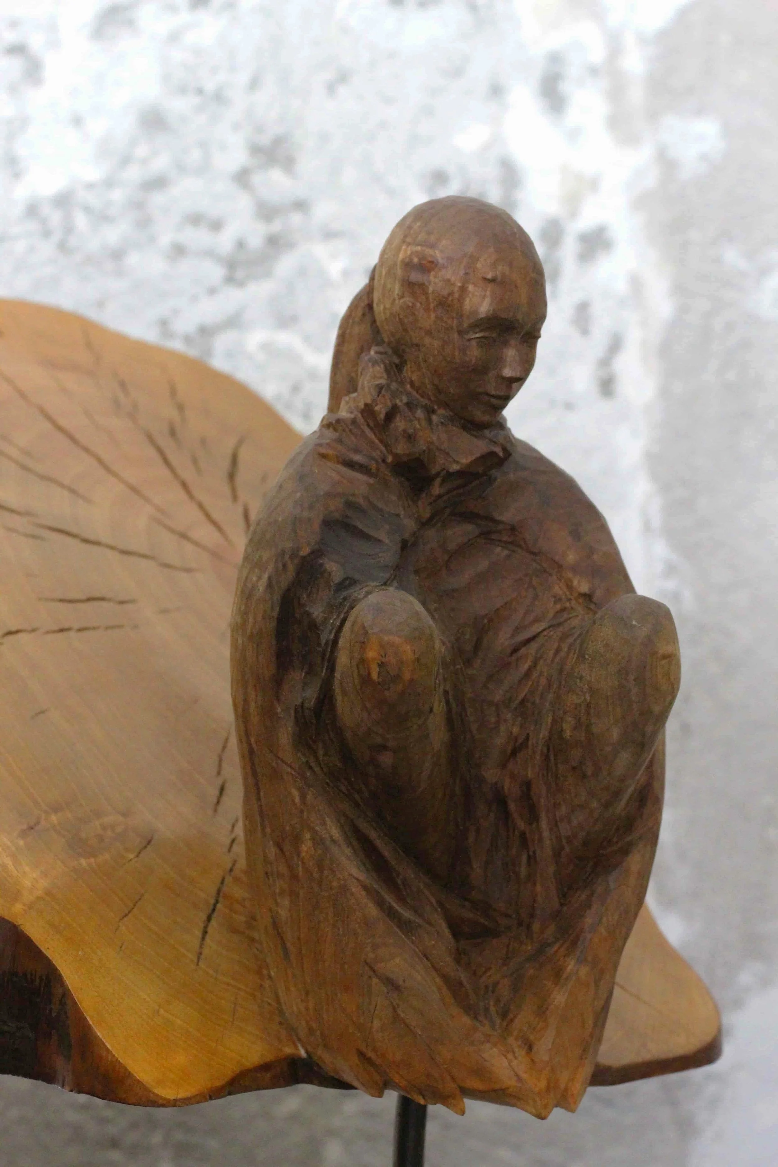 Statua di una figura femminile in legno con capigliatura liscio e occhi chiusi, seduta con le braccia incrociate, su una base di legno.