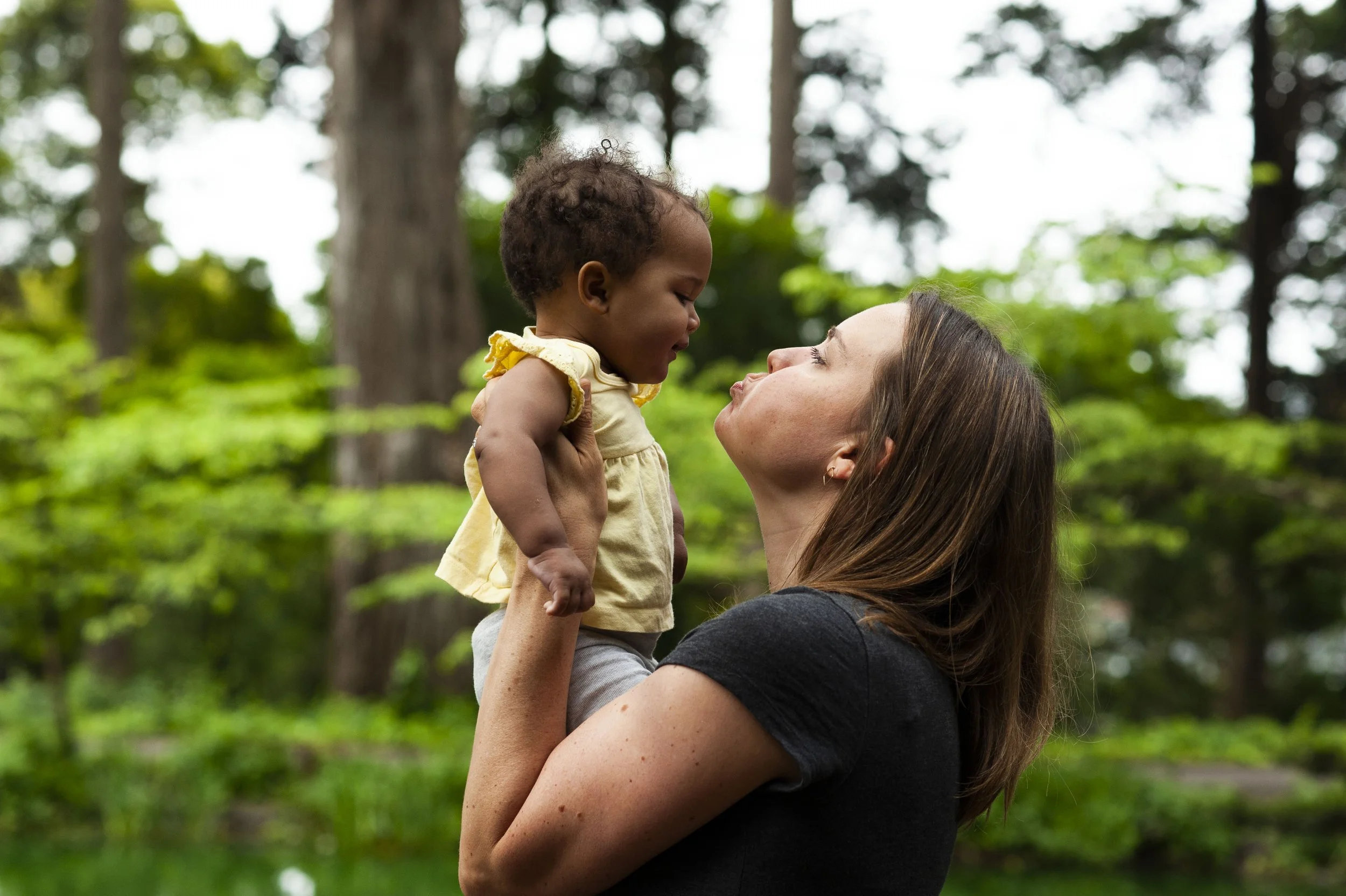 A woman holding a baby girl high in the air outdoors in a wooded area